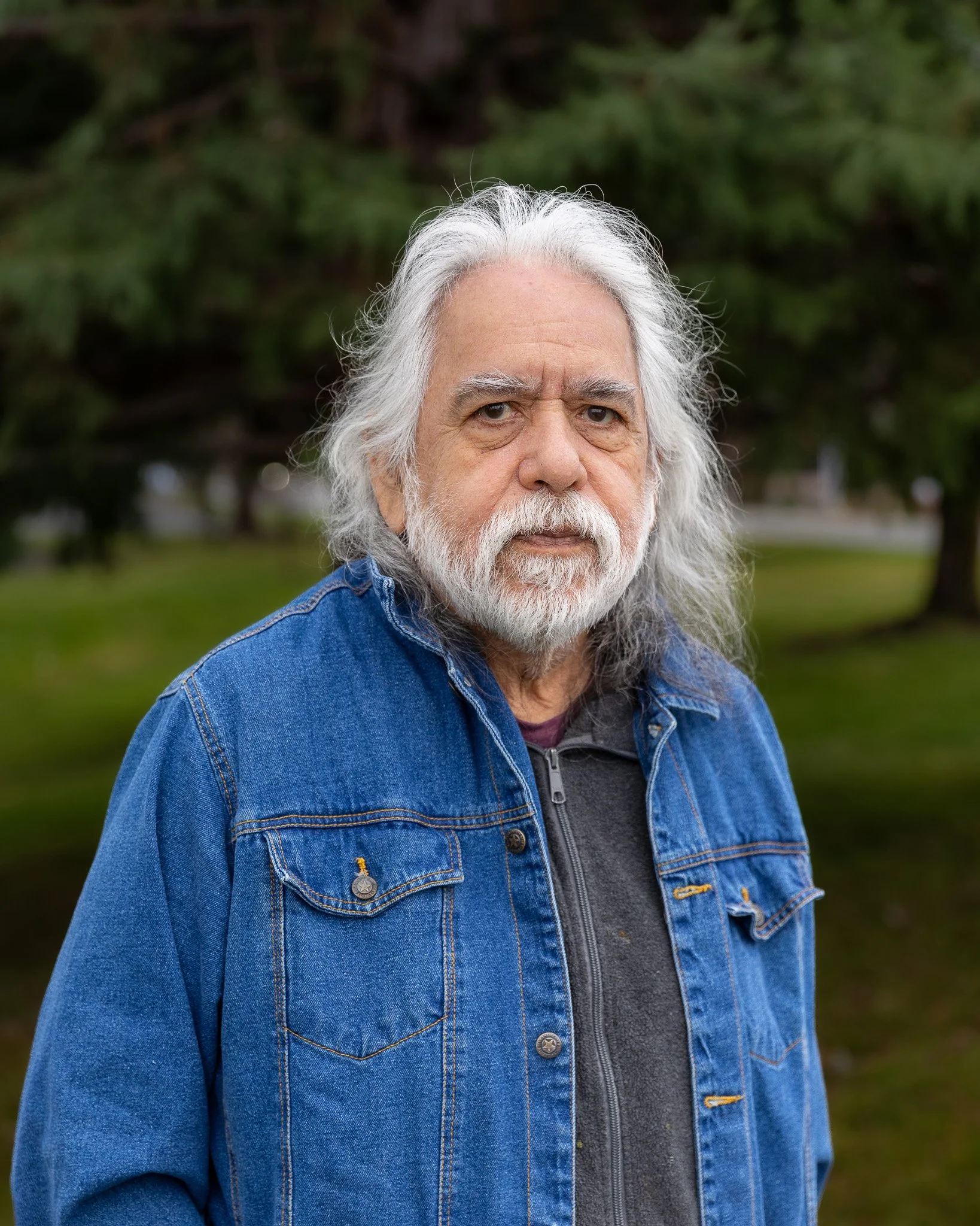 An older man with long gray hair and a beard, wearing a white shirt and a black leather vest, stands in front of a brick wall, smiling.