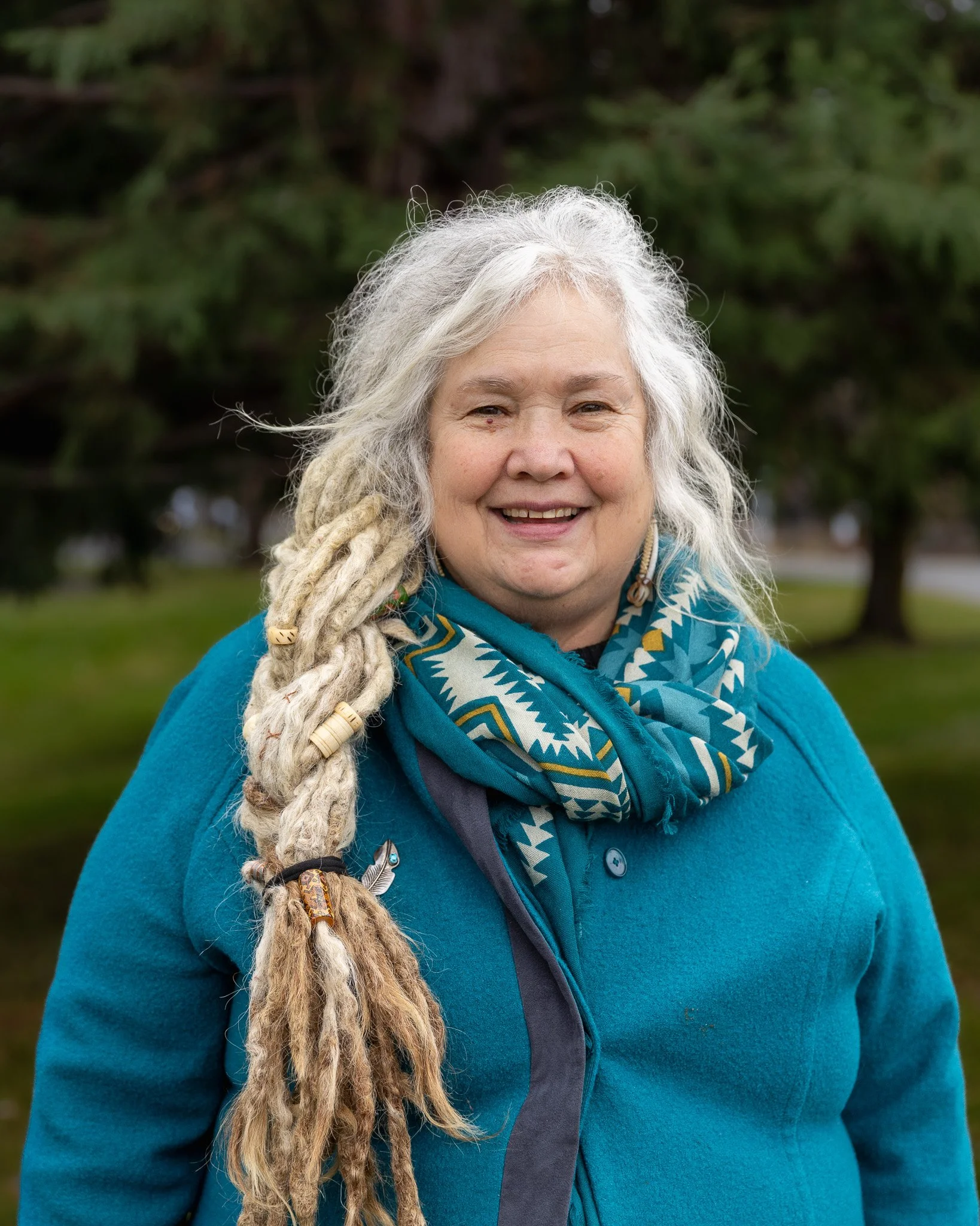 A smiling woman with long, gray dreadlocks wearing a black t-shirt that says 'DECOLONIZE ACADEMY' standing in front of a brick wall.