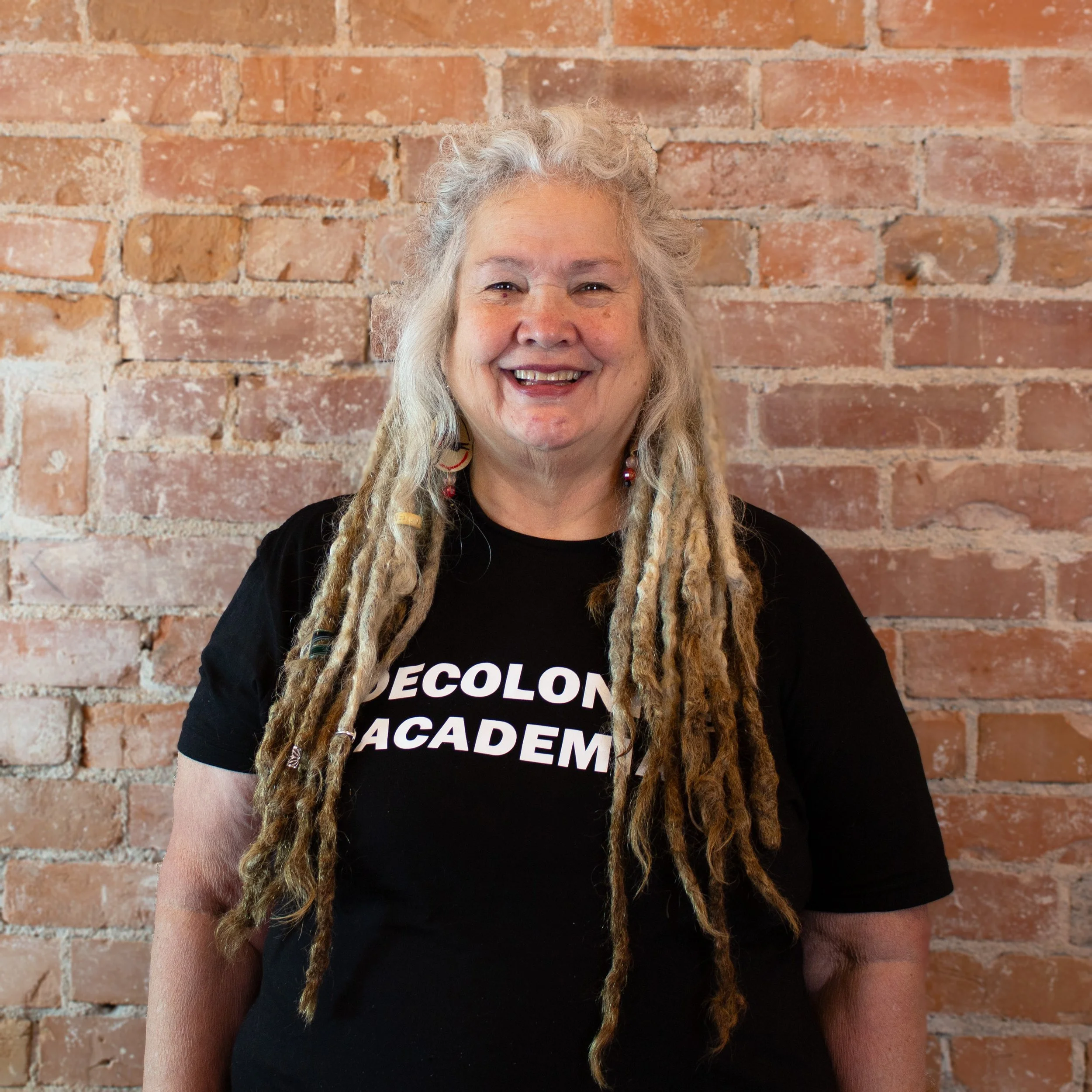 A smiling woman with long, gray dreadlocks wearing a black t-shirt that says 'DECOLONIZE ACADEMY' standing in front of a brick wall.