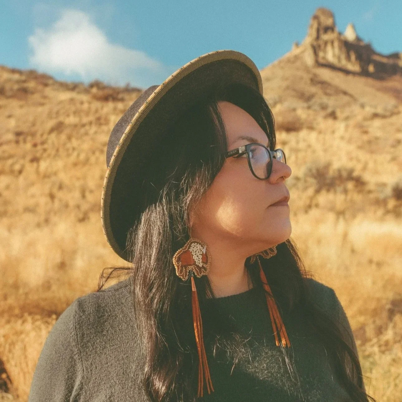 A woman with long black hair, glasses, and large floral earrings stands against a brick wall, smiling. She is wearing a colorful striped cardigan over a black T-shirt with white geometric symbols.