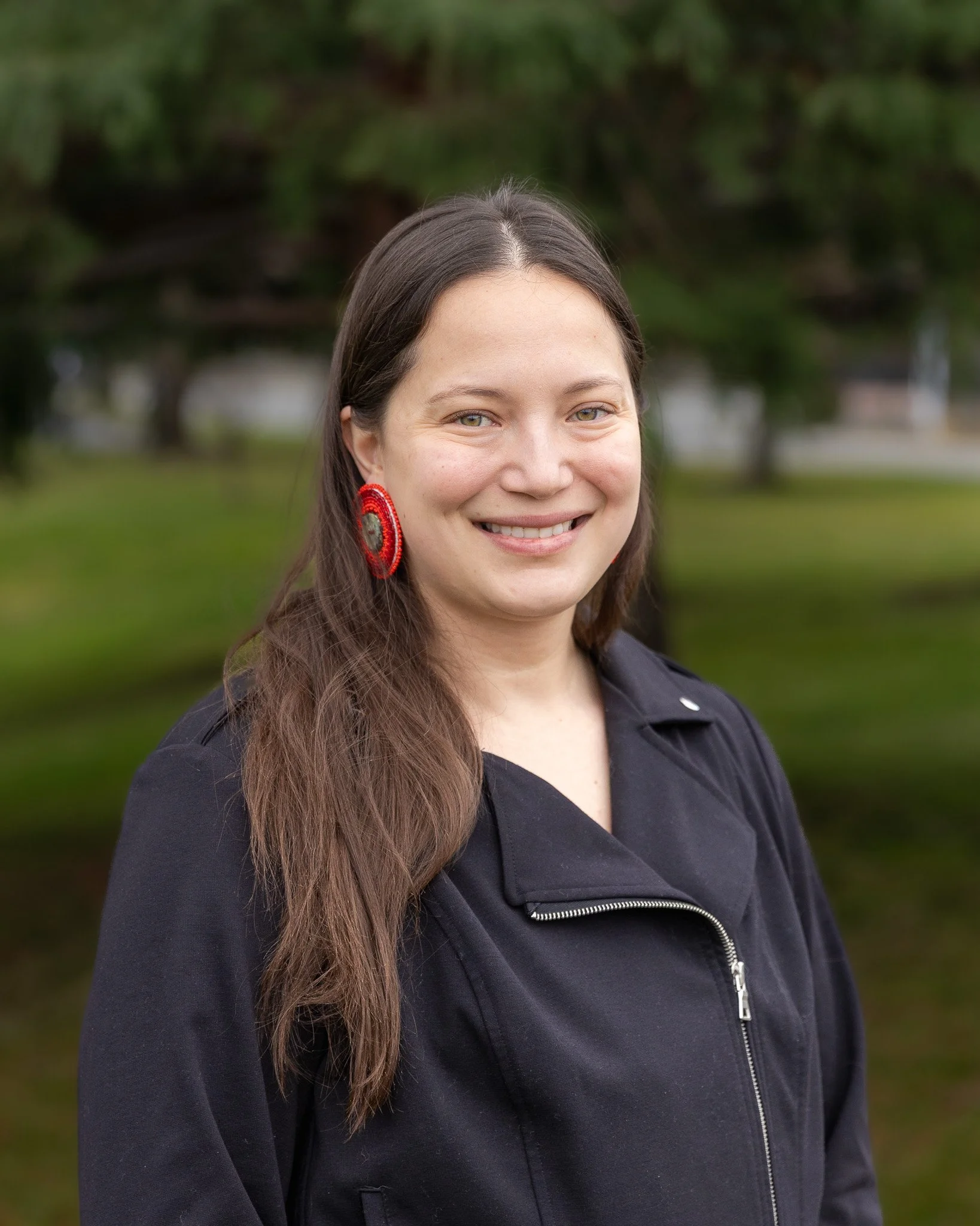 A young woman with long dark hair smiling, wearing large hoop earrings, a rectangular orange pendant necklace, a patterned shirt with black geometric designs on a tan background, and a black jacket, standing in front of a brick wall.