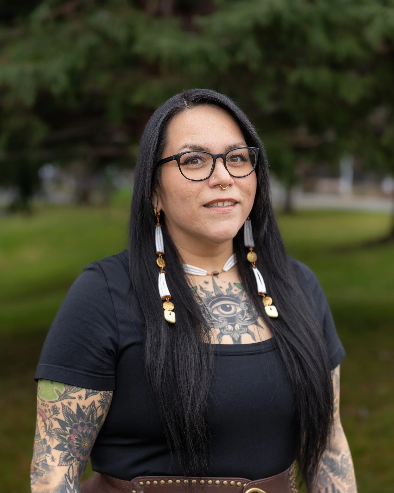 A woman with long black hair, glasses, and large floral earrings stands against a brick wall, smiling. She is wearing a colorful striped cardigan over a black T-shirt with white geometric symbols.