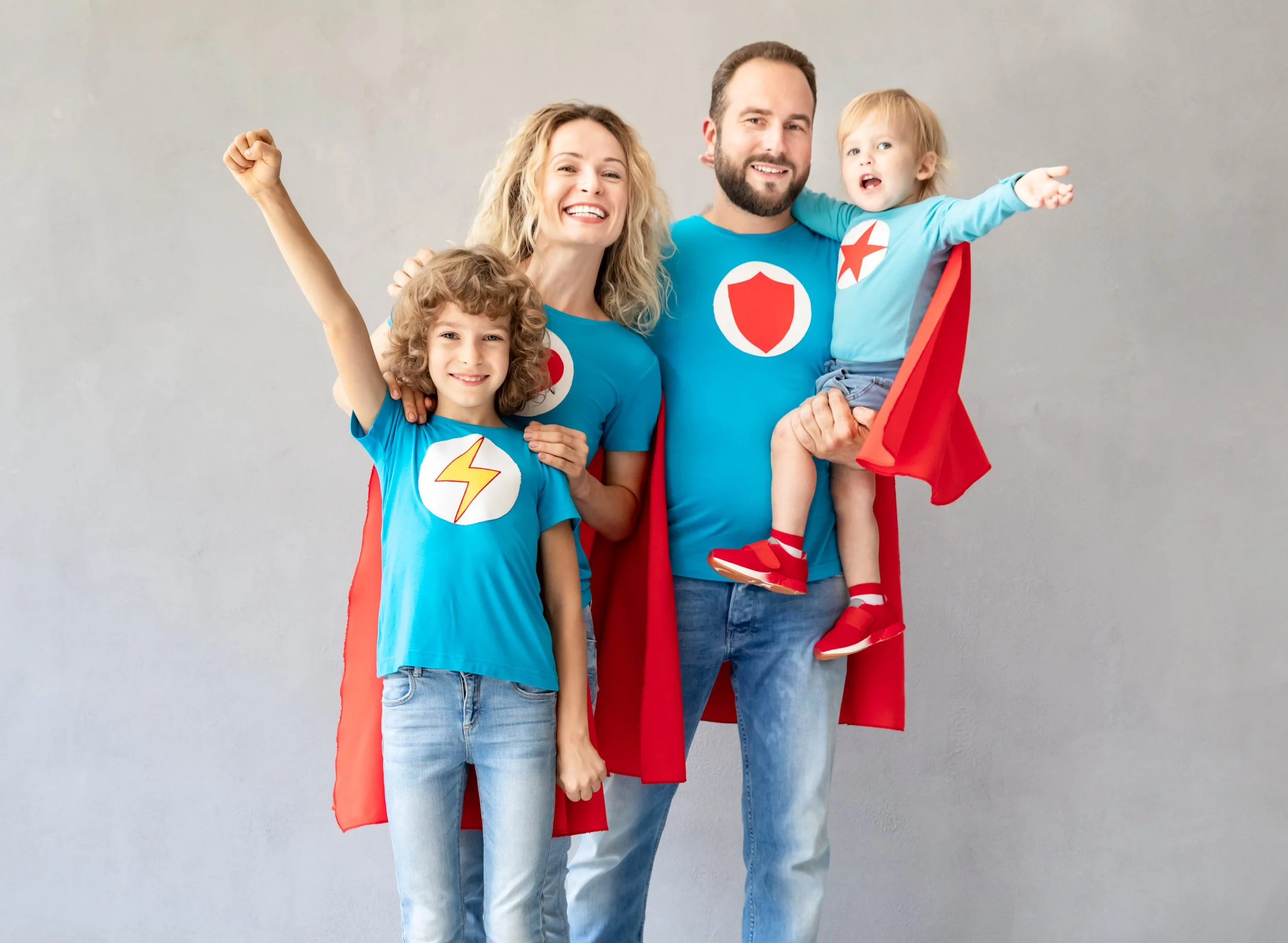 Family of four dressed as superheroes, smiling, standing against a gray wall. The mother and father wear red capes and blue shirts with superhero symbols; two children, a girl and a boy, also wear blue shirts with superhero symbols, with the girl raising her hand in a fist.