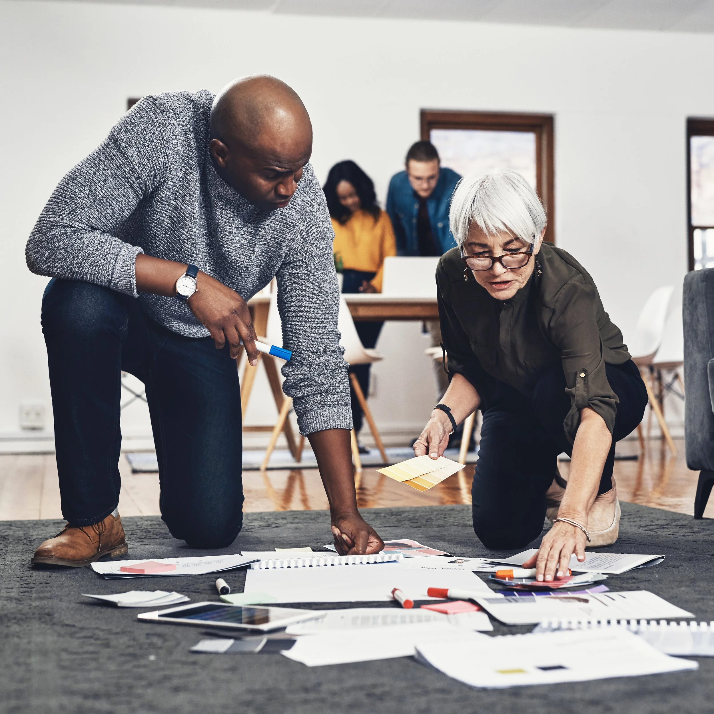 Two people kneeling on the floor and working on a project with papers, charts, and pens scattered around, while other individuals work in the background.