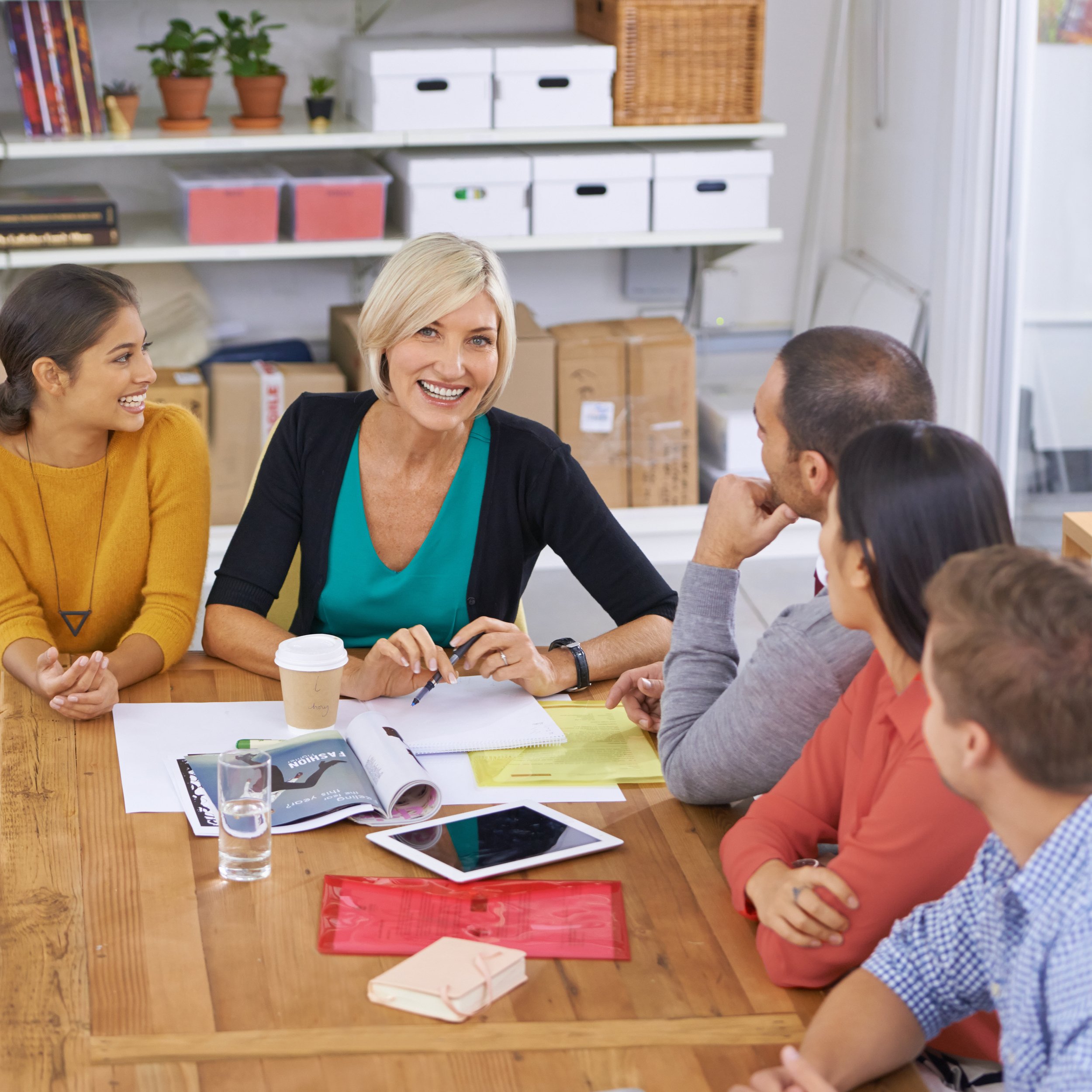A group of five people sitting around a wooden table in a meeting room, engaged in a discussion. A blonde woman in a turquoise top and black cardigan is smiling at the camera. There are notebooks, a tablet, papers, a glass of water, a coffee cup, and a pink notebook on the table.