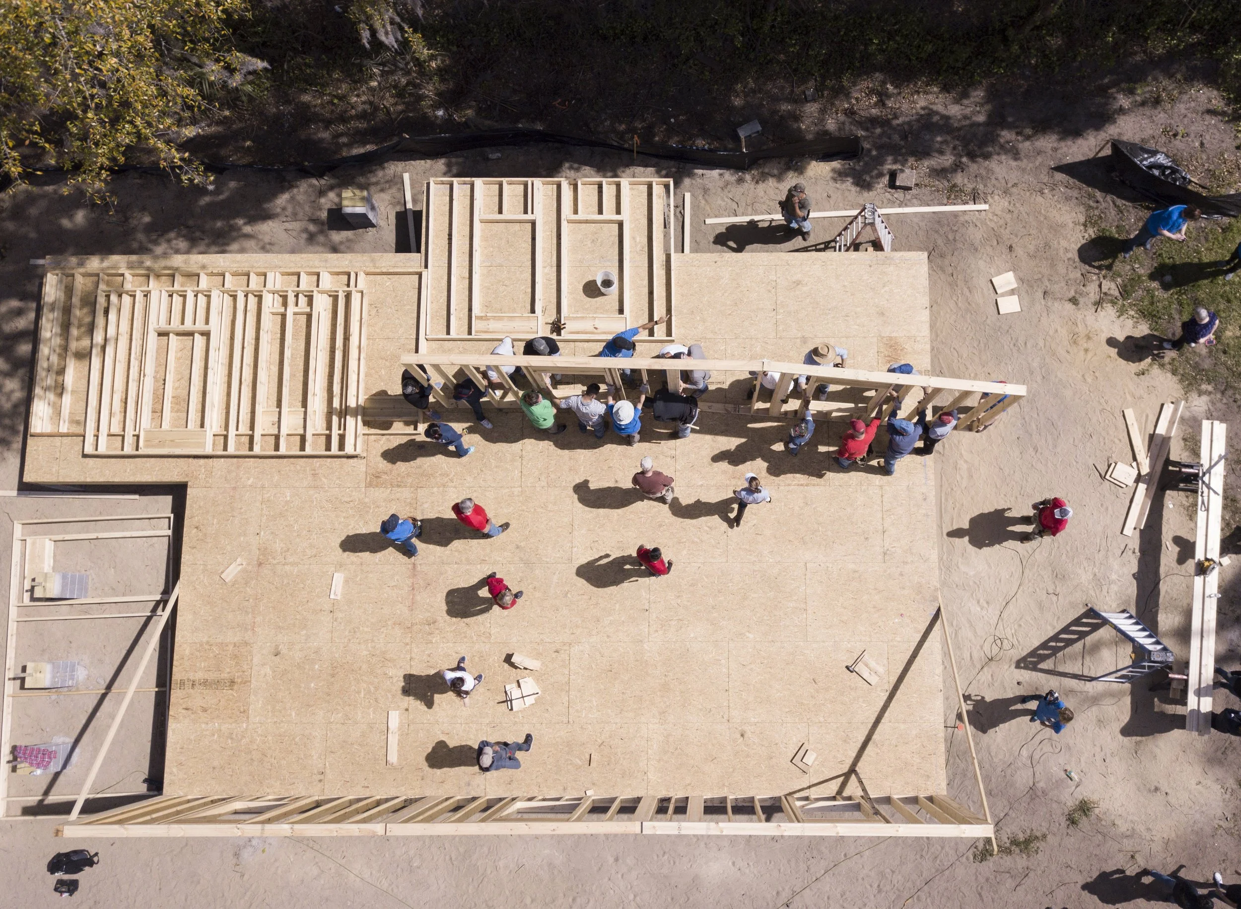 Bird's eye view of a construction site with people working on framing a building. The partially built wooden structure is surrounded by sandy ground, with some trees at the top edge of the image.