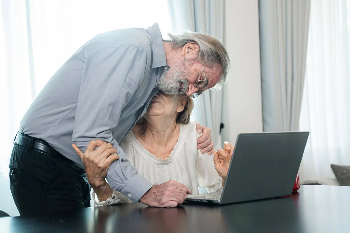 An elderly man leaning over an elderly woman sitting at a table with a laptop, showing affection and hugging her.
