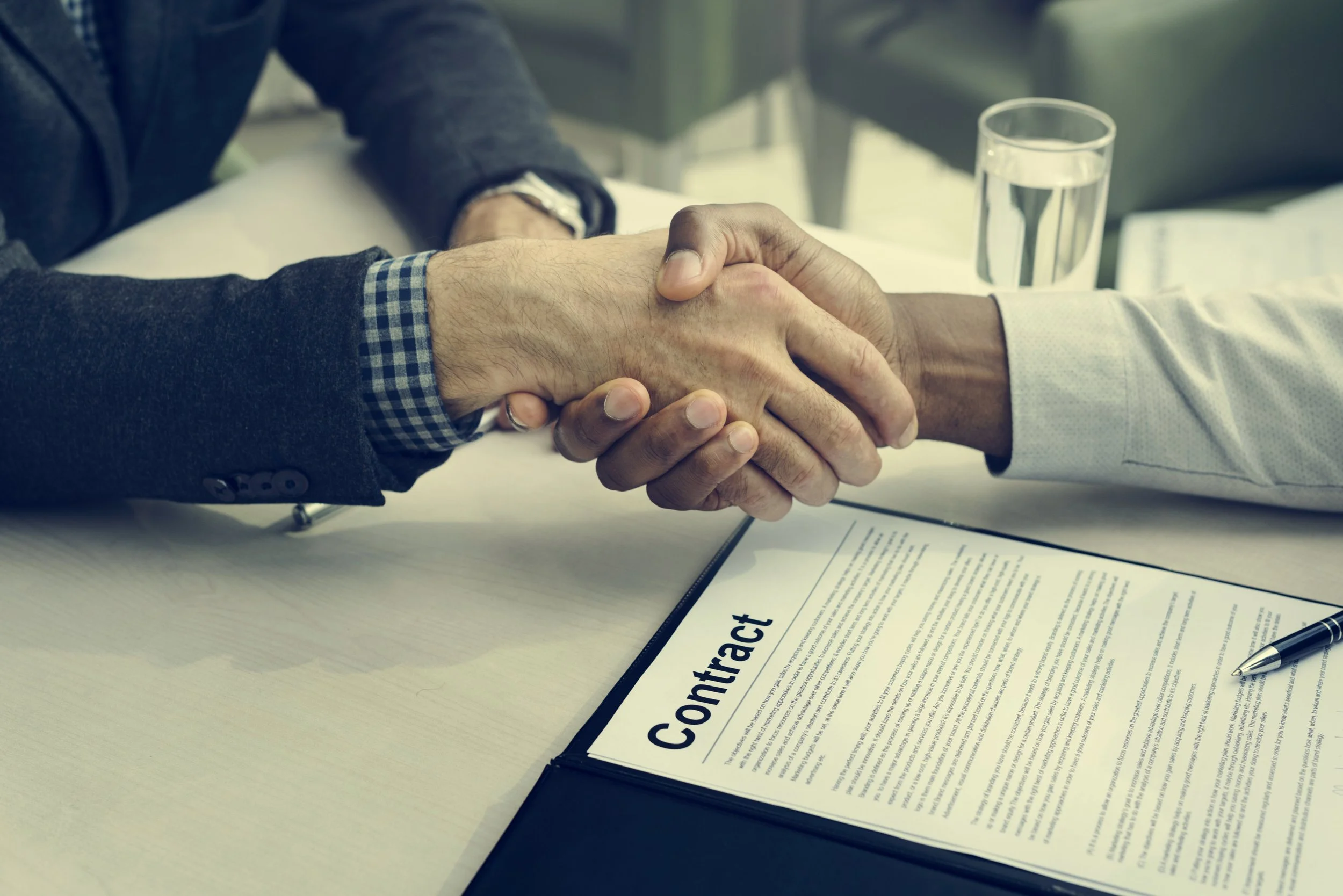 Two individuals shaking hands over a contract on a table, with a pen and a glass of water nearby.