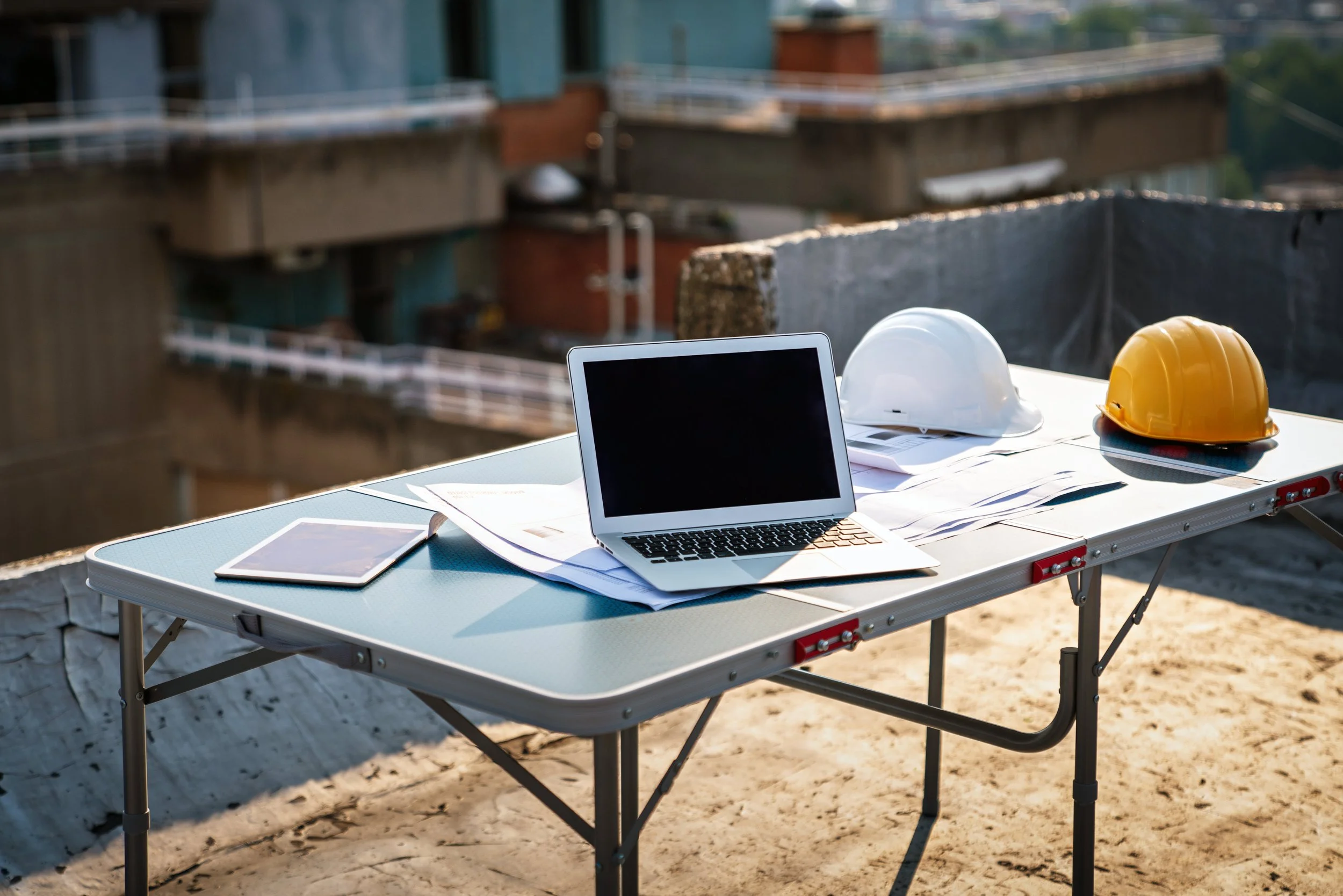 Construction site desk with a laptop, a tablet, blueprints, and two safety helmets on a portable table.