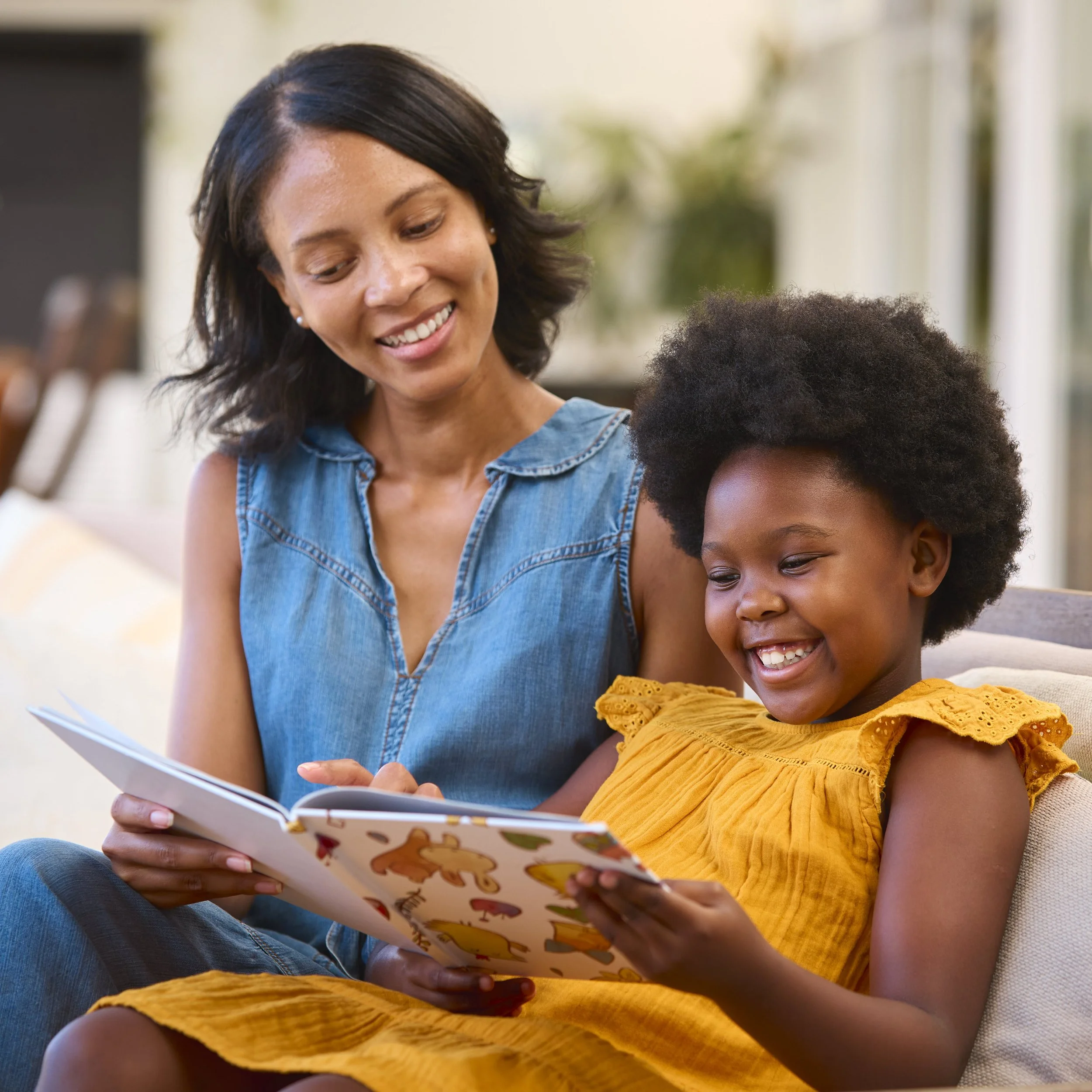 A woman and a young girl are sitting together on a couch, reading a children's book and smiling.