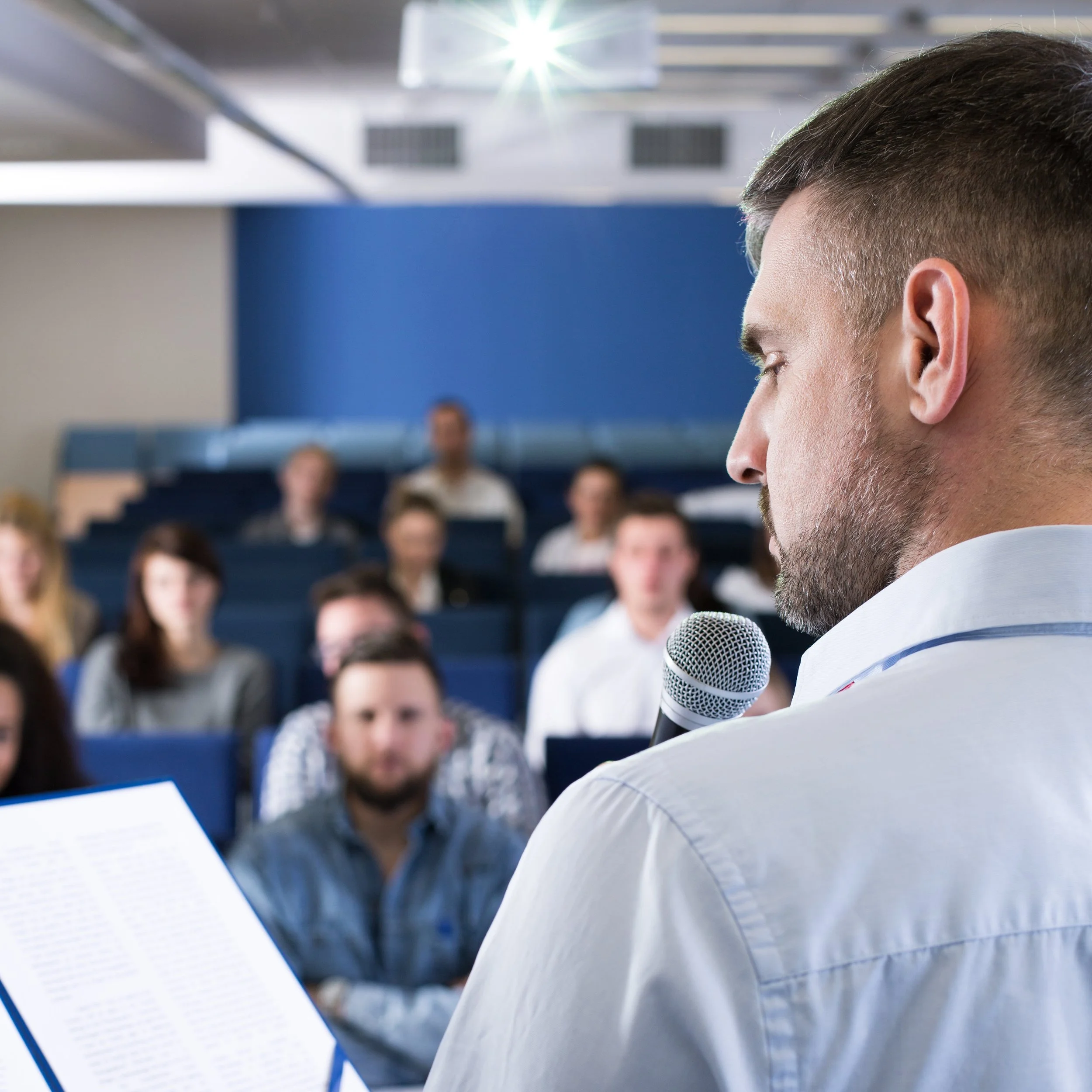 A man with a beard is holding a microphone and a sheet of paper, speaking to an audience seated in a lecture hall.