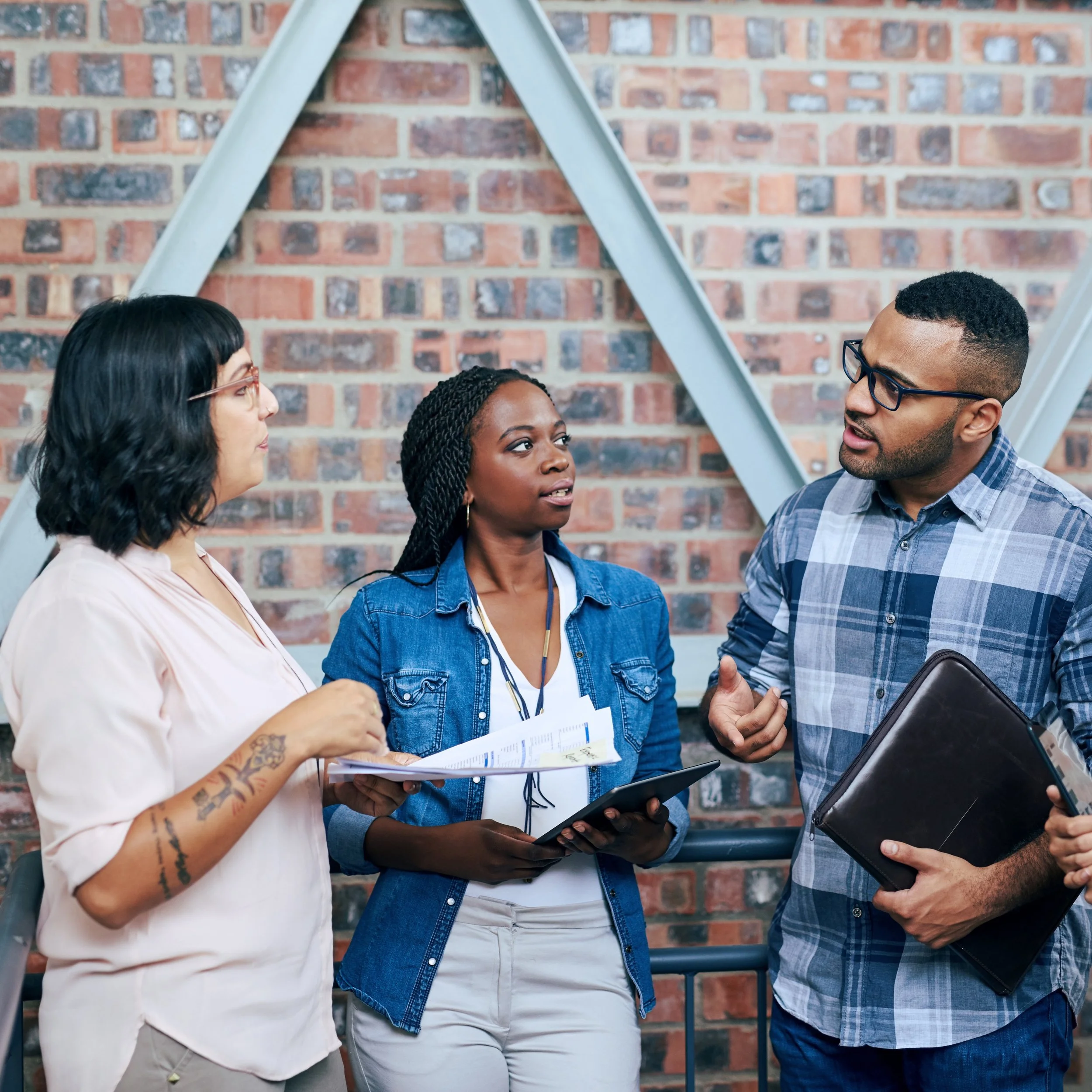 Three diverse young professionals engaged in a discussion indoors, with a brick wall and structural beams behind them.