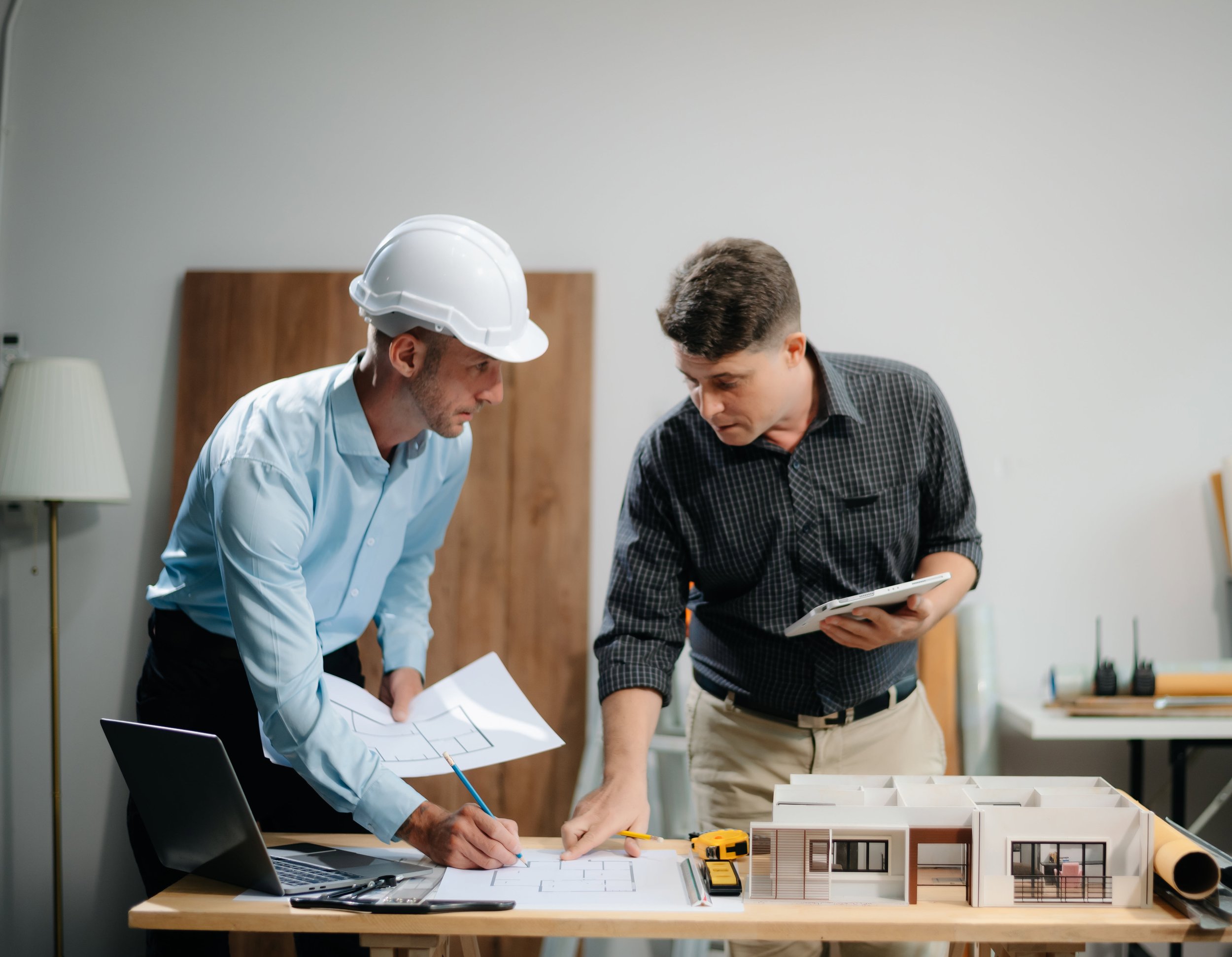 Two men, one wearing a white safety helmet, discussing architectural plans and models on a table in an office or studio.