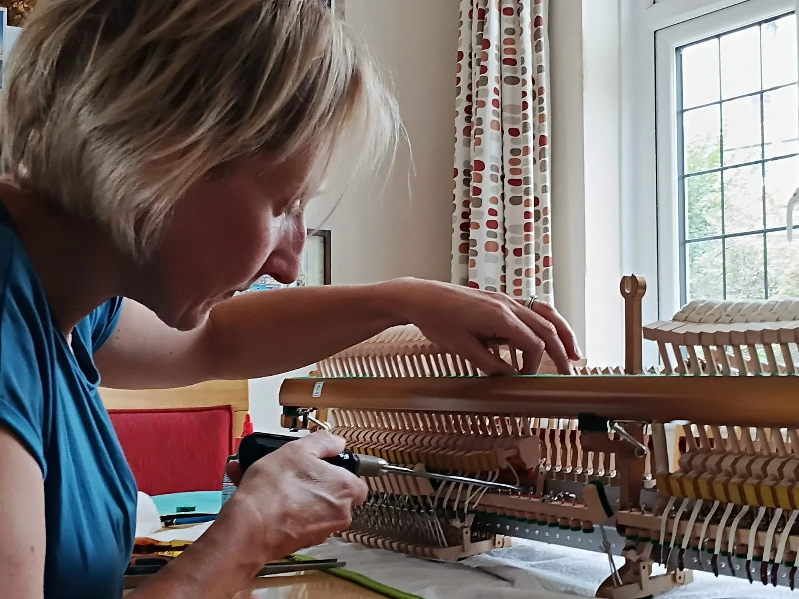 A woman working on the internal components of a piano, using a screwdriver, with a window and curtains in the background.