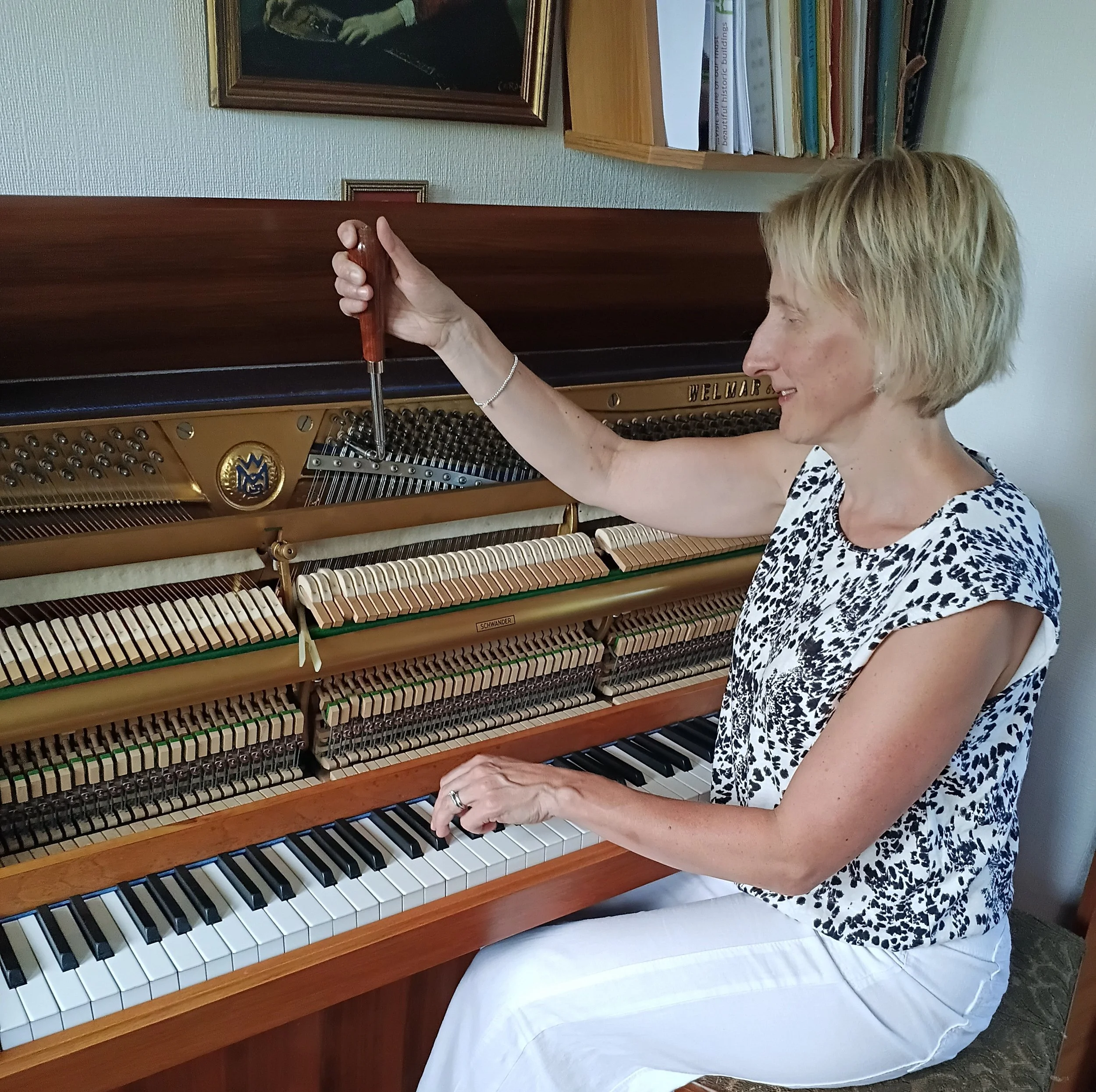 A female piano tuner with short blonde hair, wearing a black and white patterned sleeveless top and white trousers, is sat at an upright piano and holding a tuning hammer.