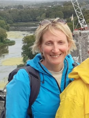 Smiling woman outdoors near a river with trees, a bridge and a crane in the background.