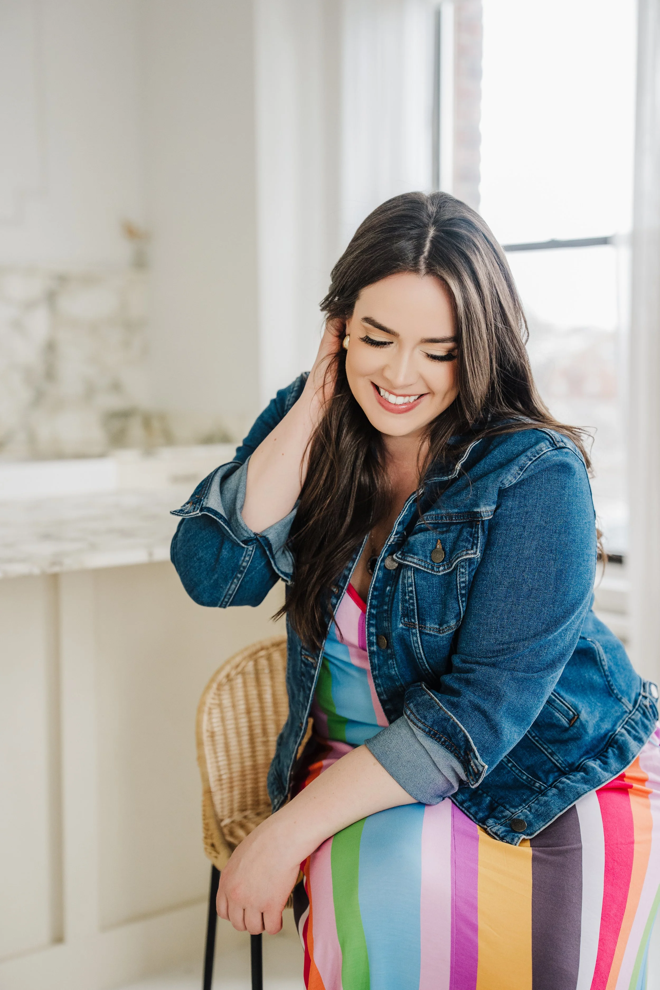 A woman with long brown hair, wearing a rainbow-colored dress and denim jacket, smiling and holding her head in a brightly lit room with large windows.