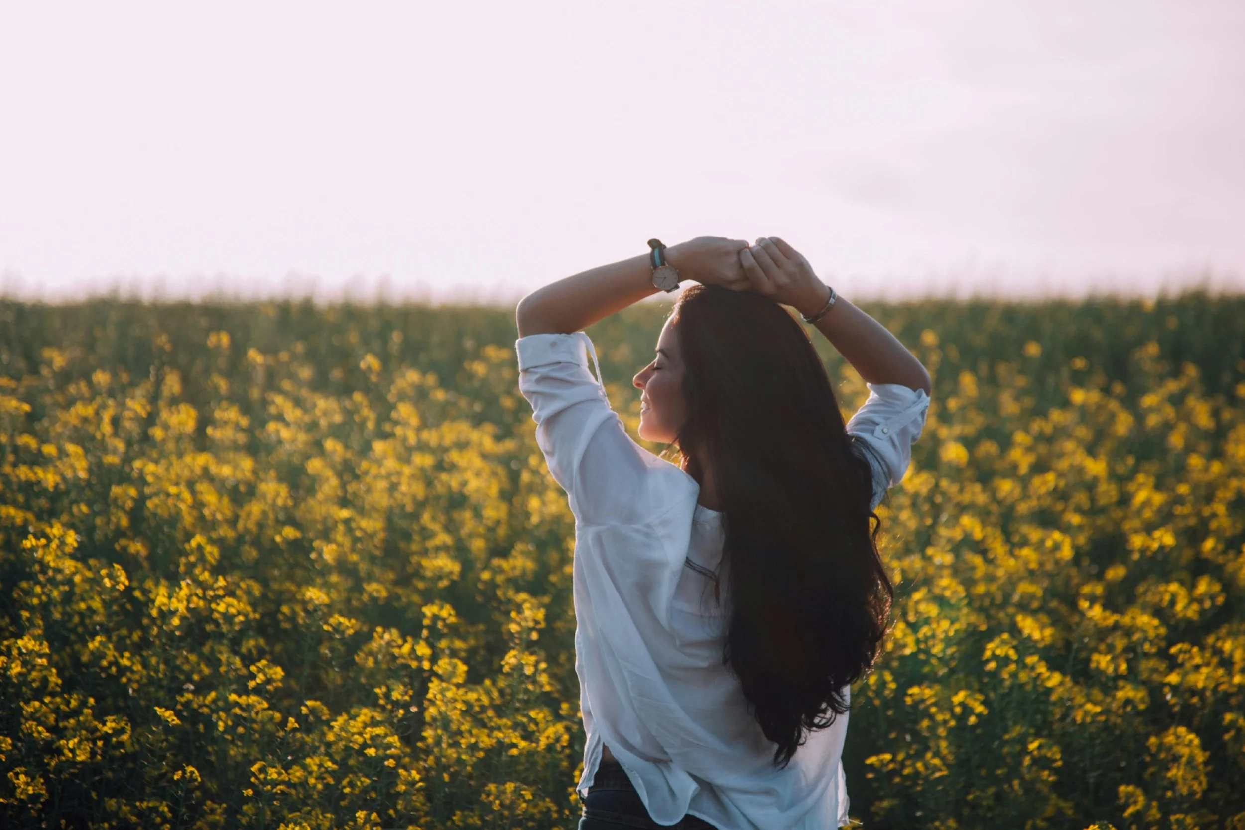 Woman in a white shirt standing in a field of yellow flowers at sunset, with her hands resting on her head and her eyes closed.
