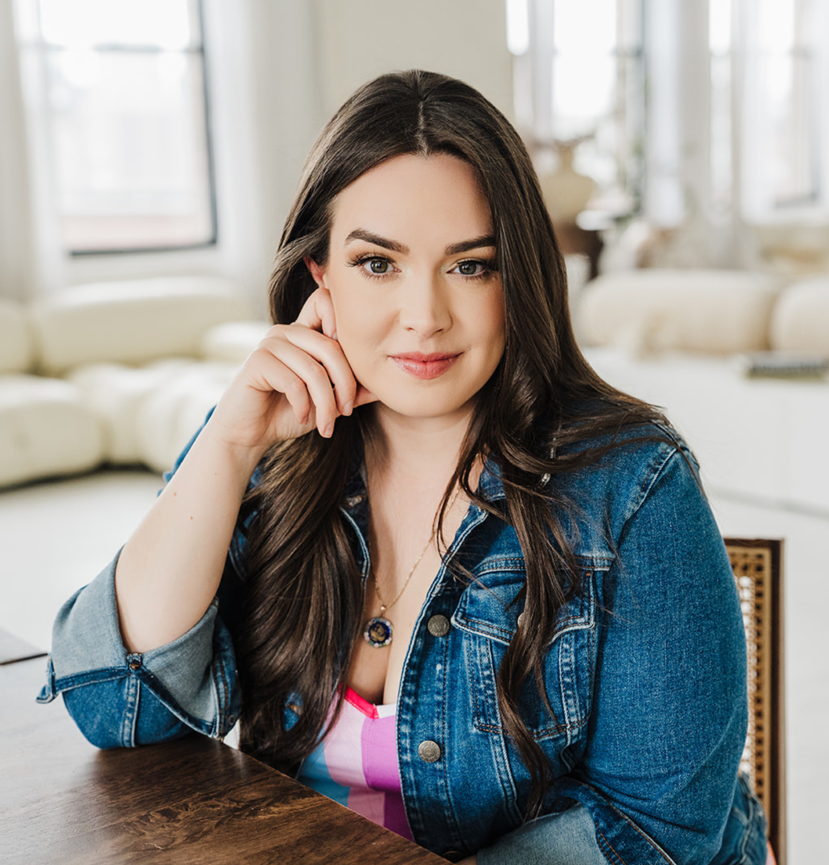 A woman with long dark hair and fair skin sitting at a wooden table in a bright, well-lit room with large windows and white furniture, wearing a denim jacket and a necklace.