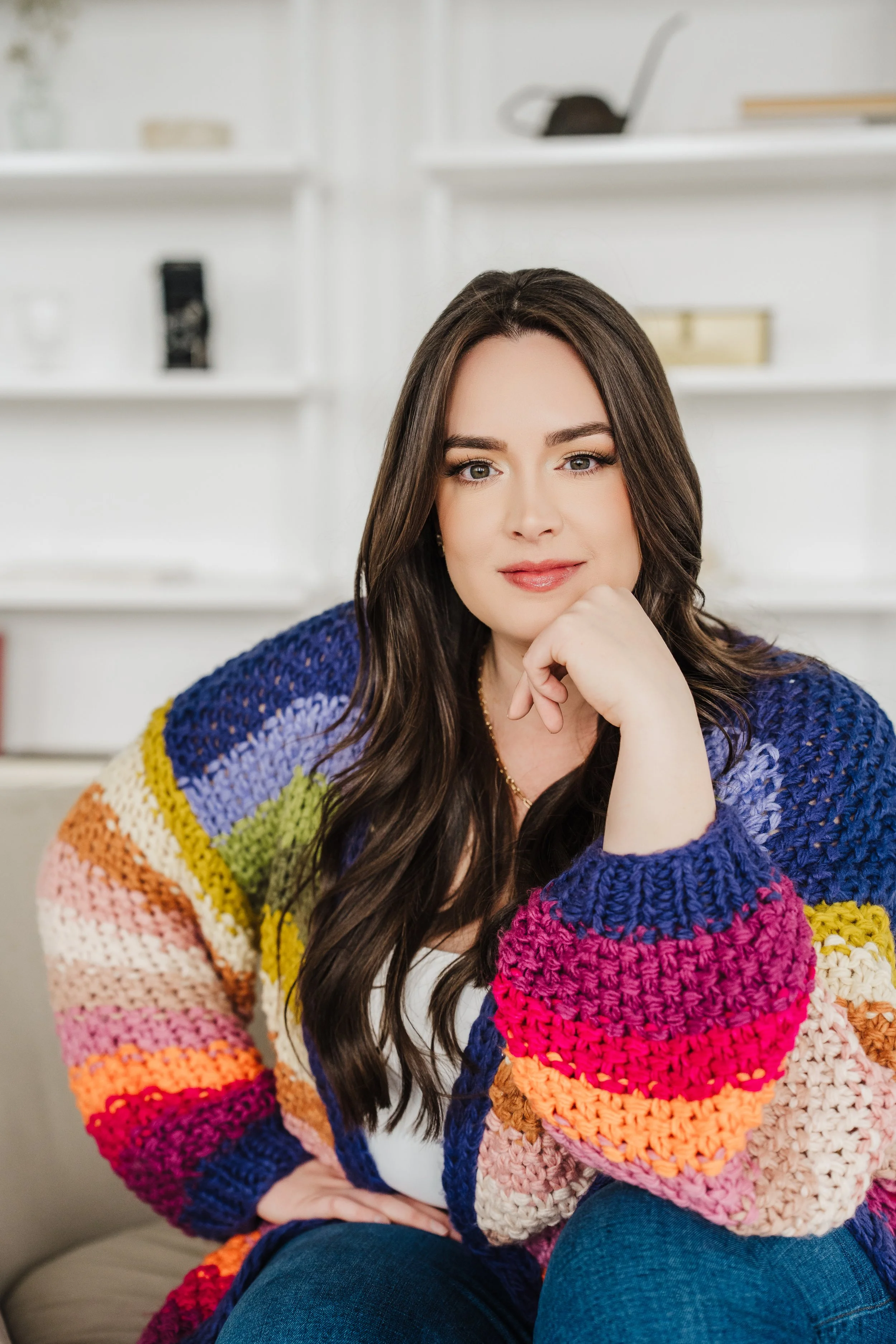 A woman with long brown hair, wearing a multicolored knitted sweater, sitting on a beige couch in a bright room with white shelves in the background.