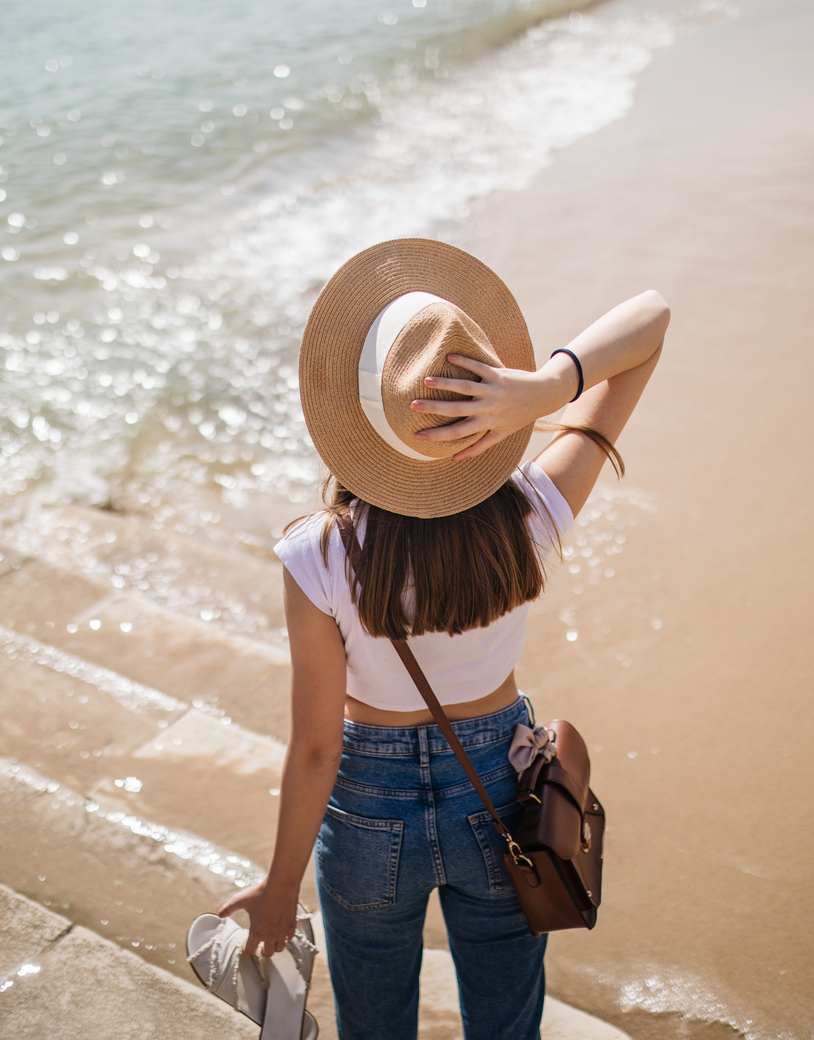 A woman in a white crop top and blue jeans holding sandals, wearing a wide-brimmed straw hat, standing on a sandy beach near the water.