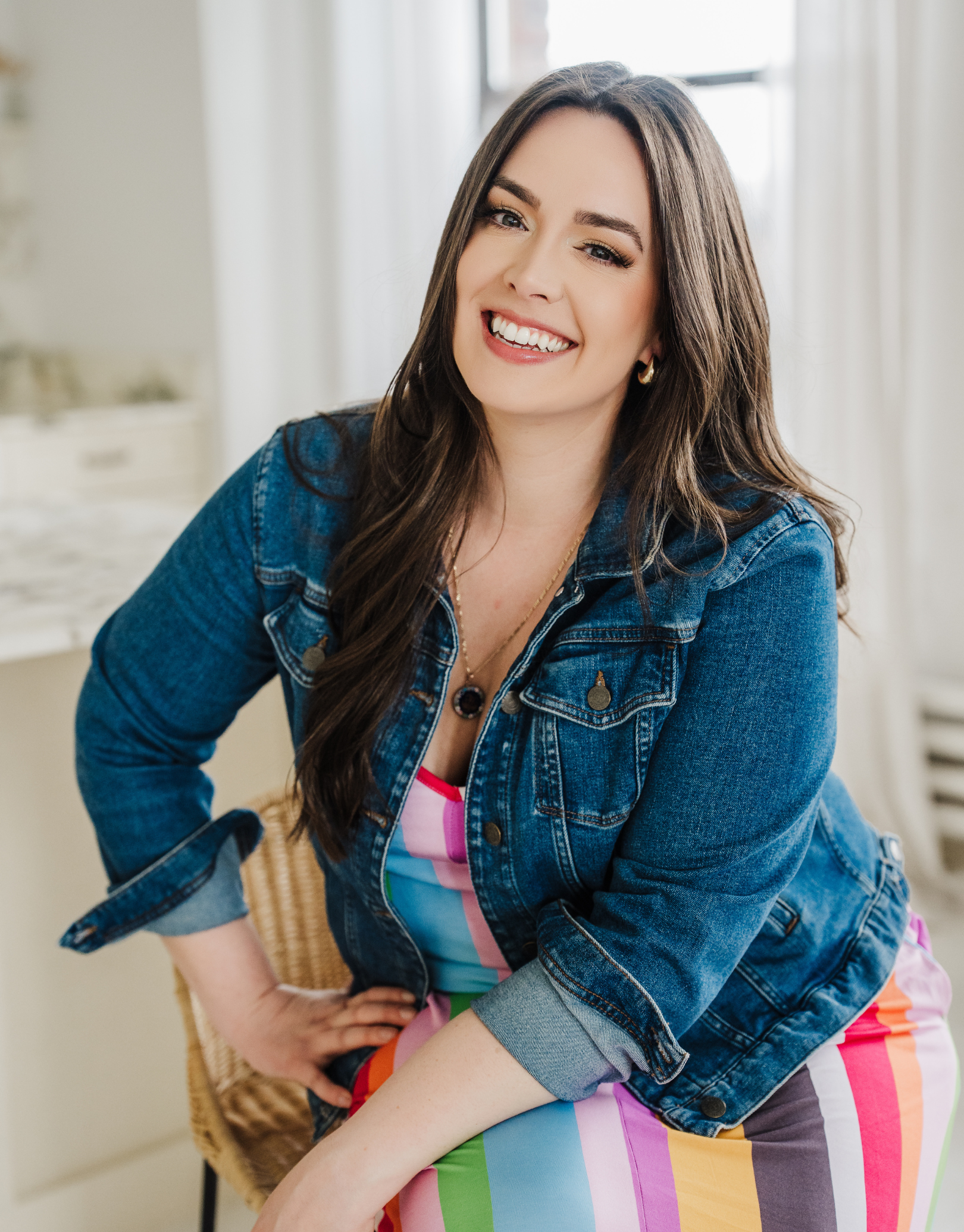 A woman with long brown hair wearing a denim jacket and a colorful rainbow-striped dress, smiling and leaning forward.