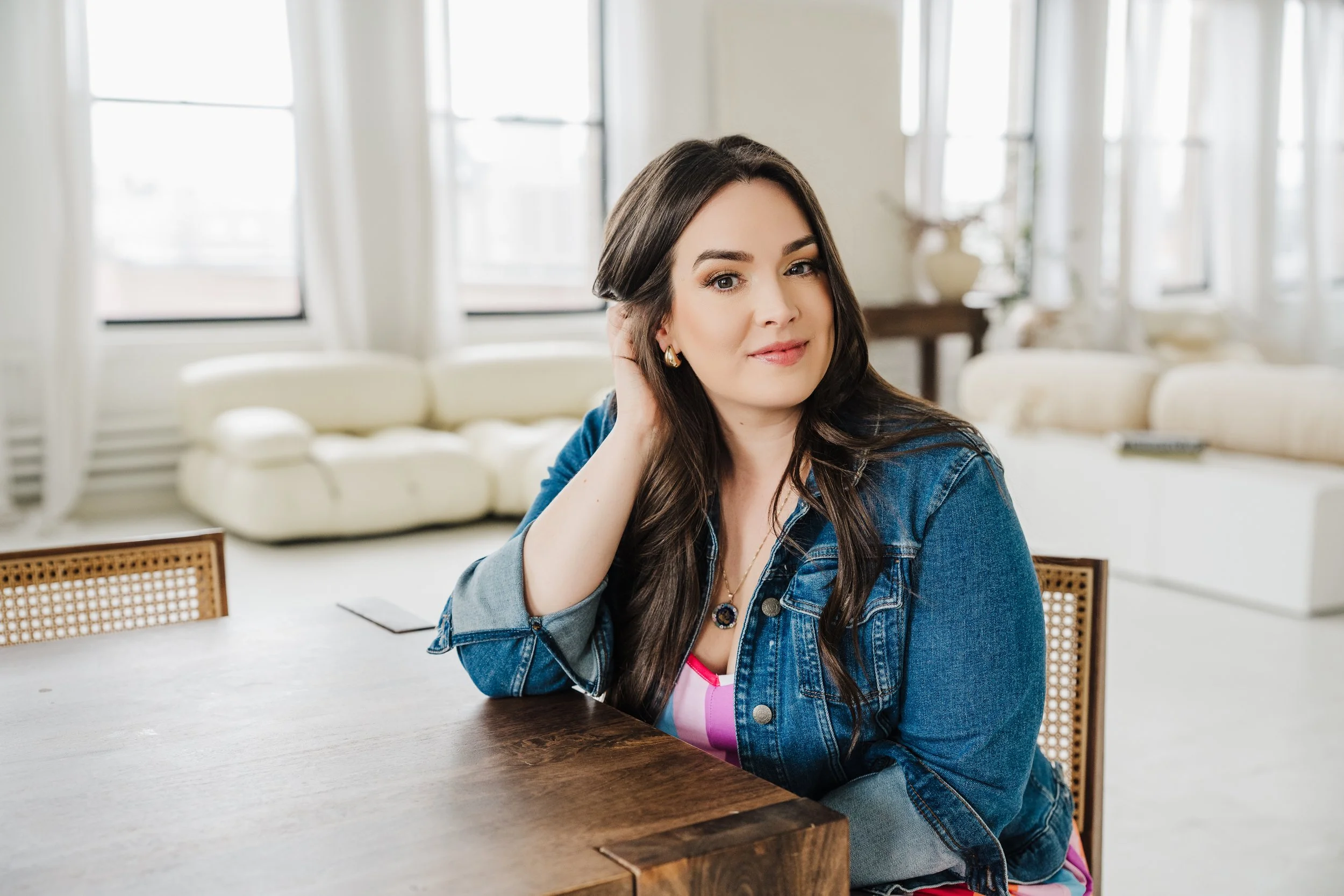 A woman with long dark hair wearing a denim jacket, sitting at a wooden table in a bright living room with large windows and white furniture.