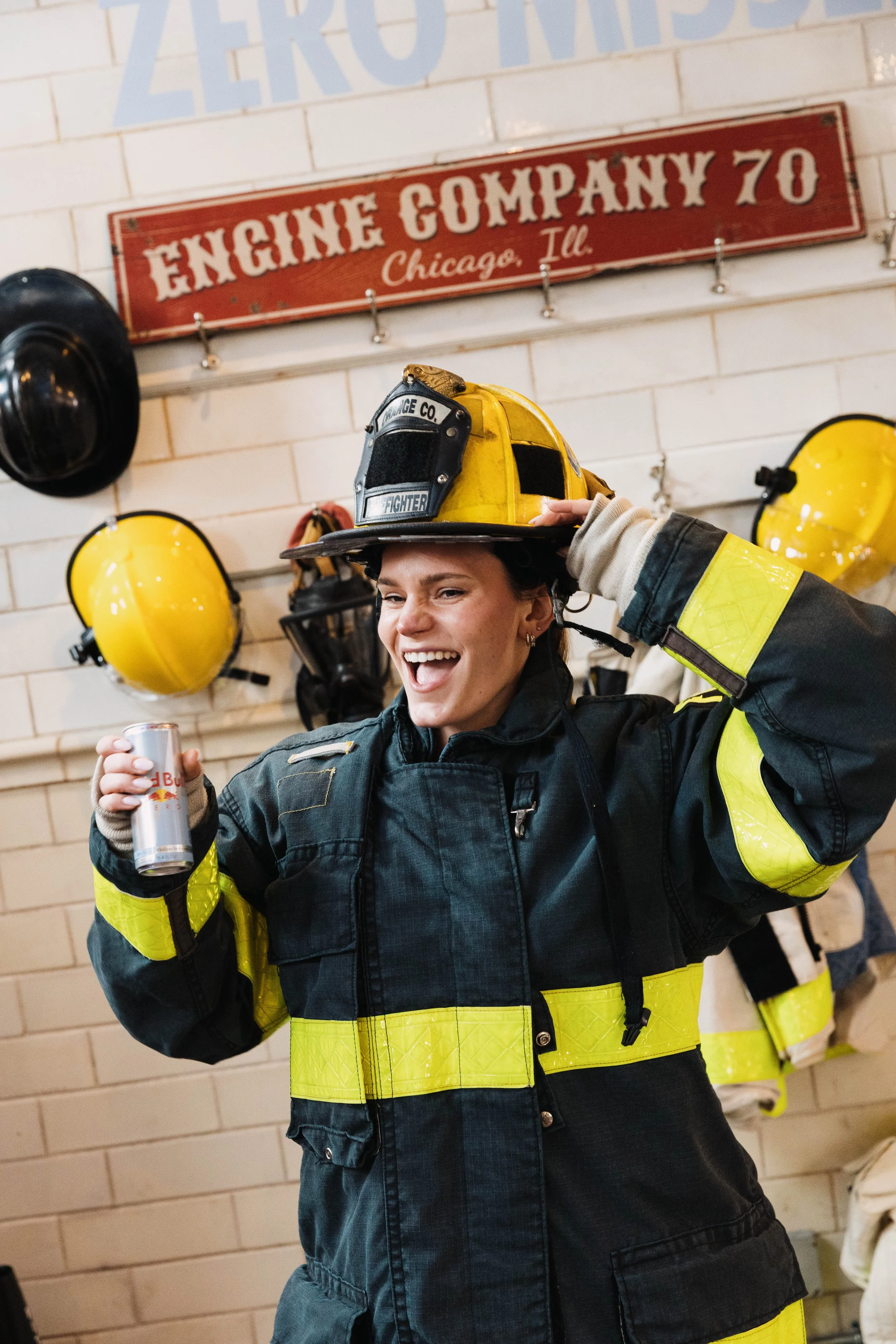 A smiling woman in firefighter gear holding a Red Bull can, standing in front of a wall with firefighter helmets and gear hanging, and a sign that reads 'Engine Company 70 Chicago Ill'.