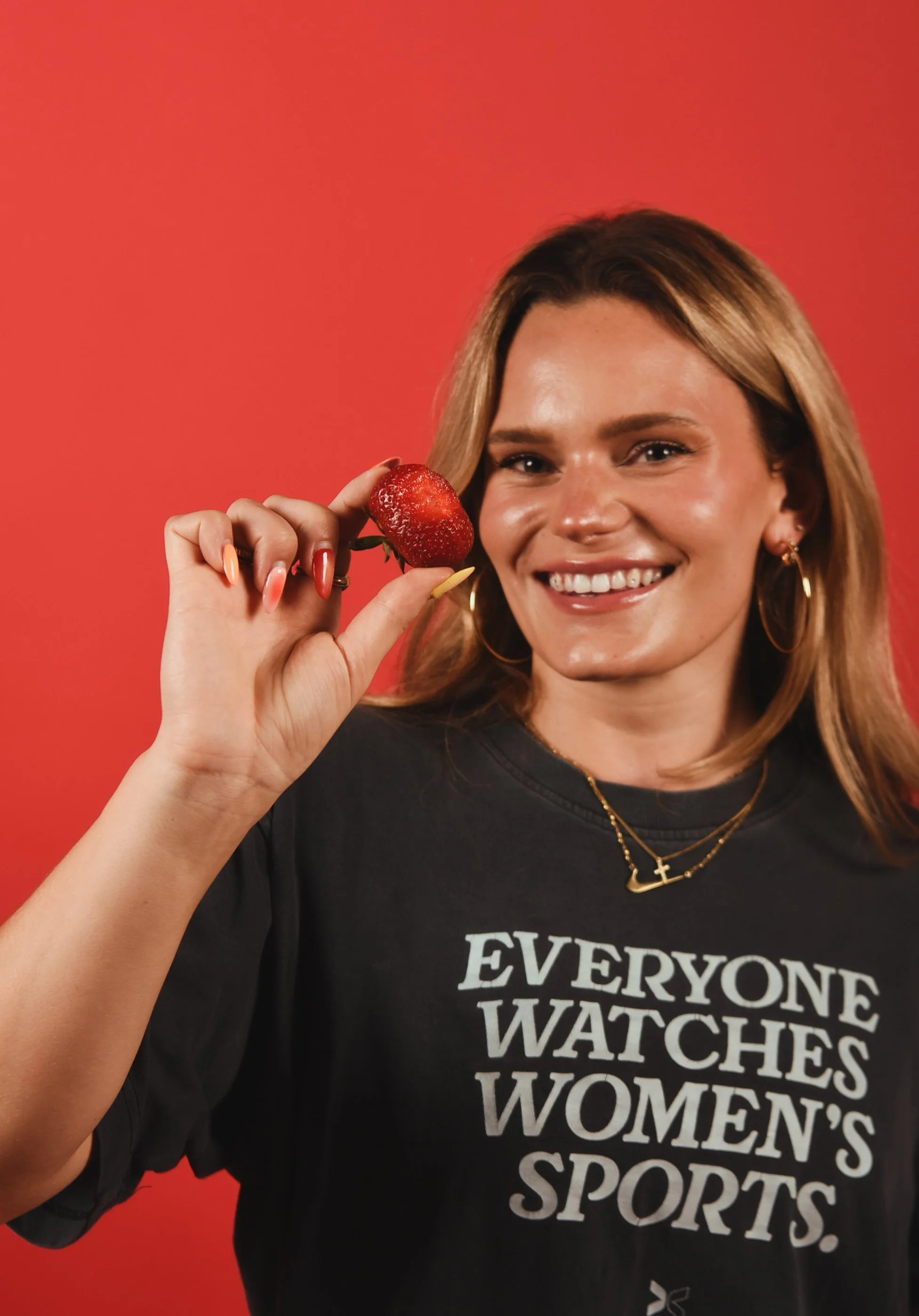 A smiling woman with blonde hair wearing a black t-shirt that says 'Everyone Watches Women's Sports,' holding a strawberry near her face against a red background.