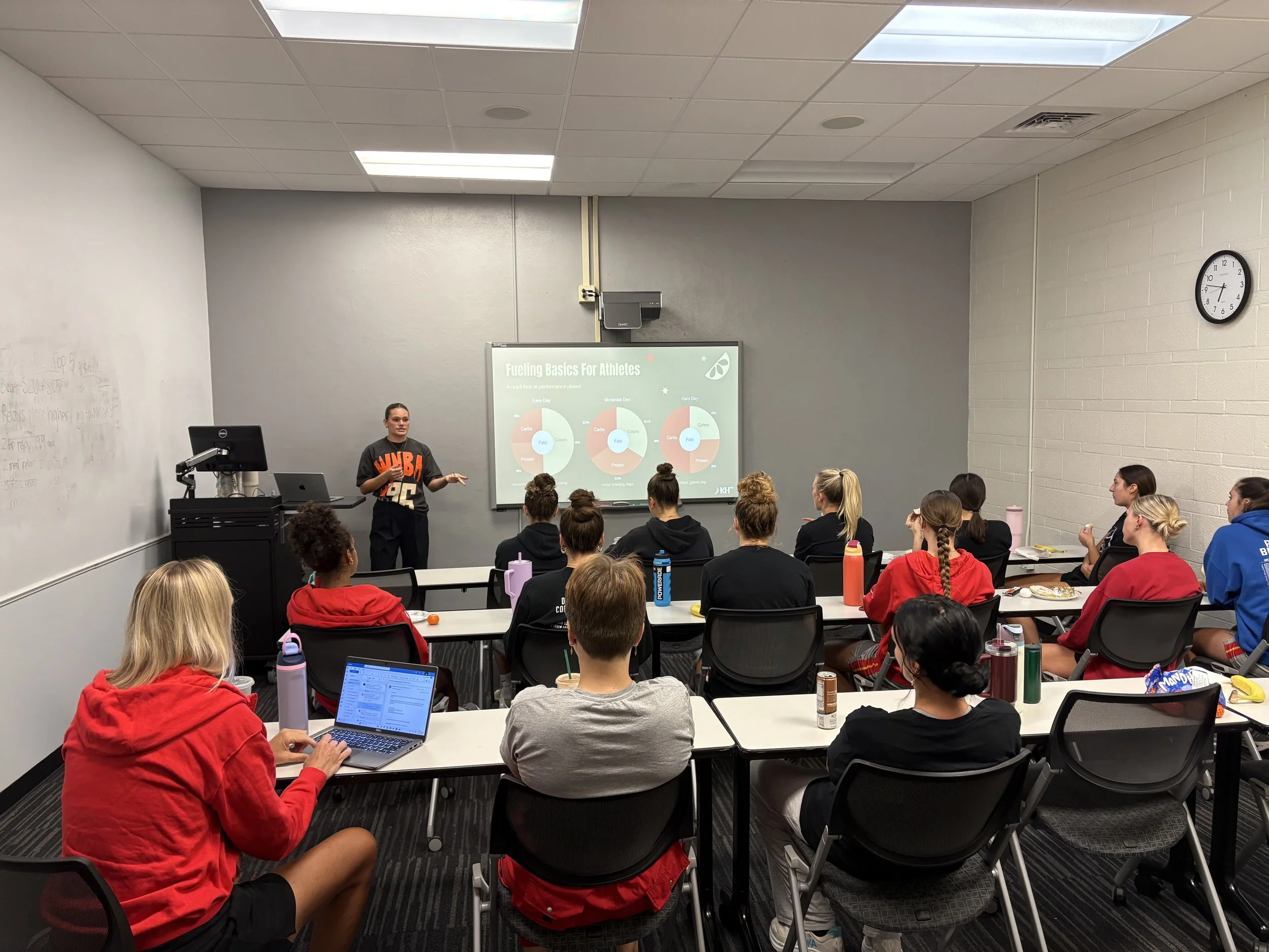 A classroom with a presenter at the front, giving a presentation titled 'Fueling Basics For Athletes,' with students seated at desks, some taking notes on laptops and others listening attentively.