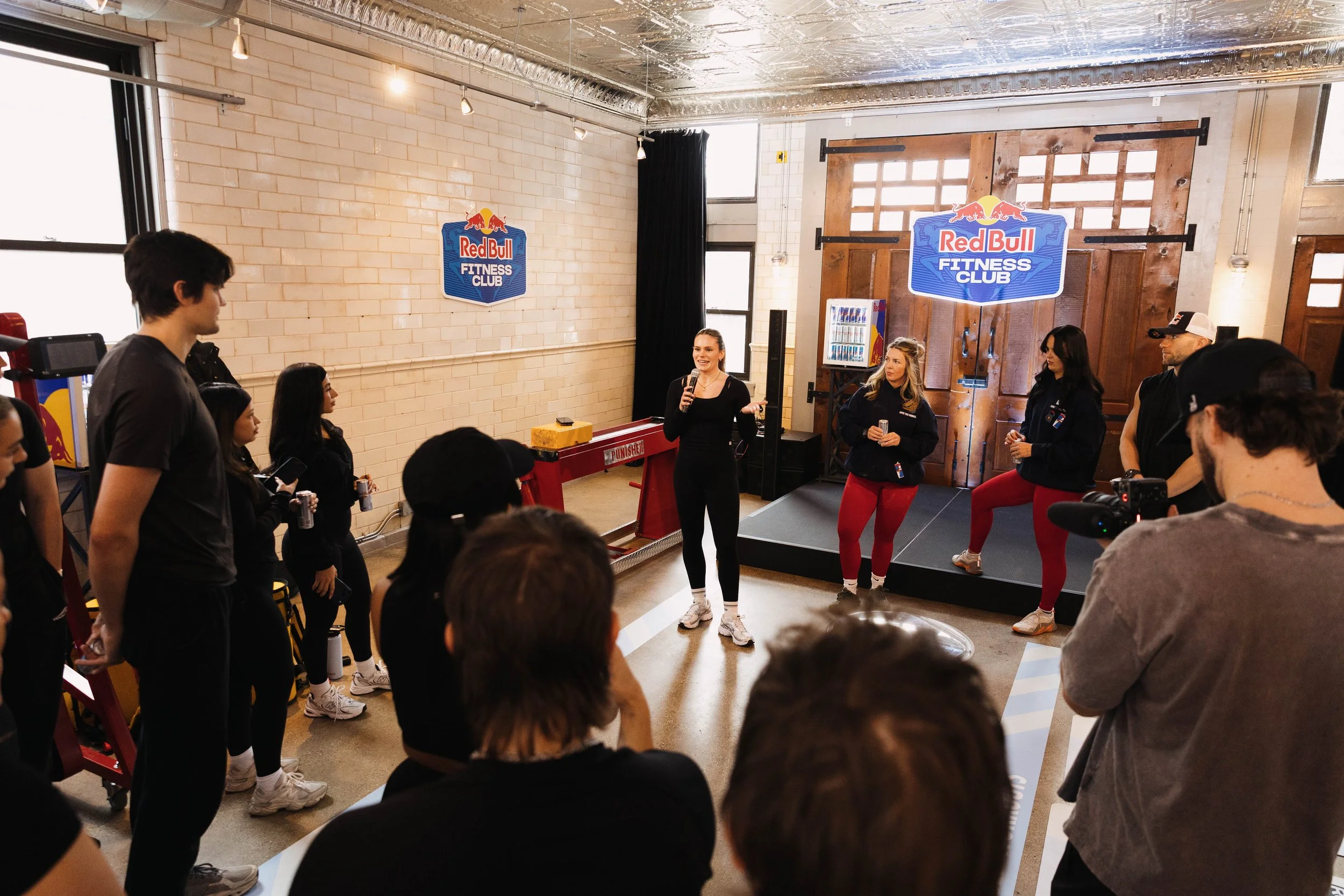 A woman speaking on a microphone in front of a group of people in a fitness club with a Red Bull logo and fitness club signage on the wall.