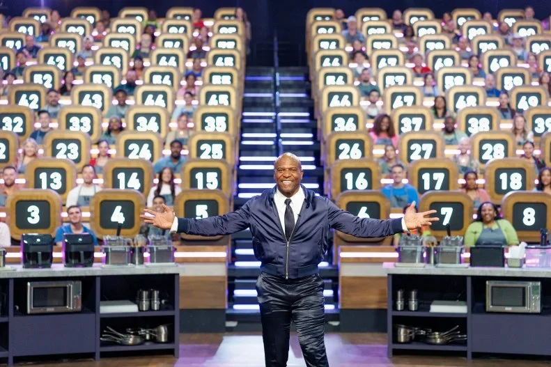 A game show host standing in the center of a studio with a large audience, with numbered seats behind him and game booths in front.
