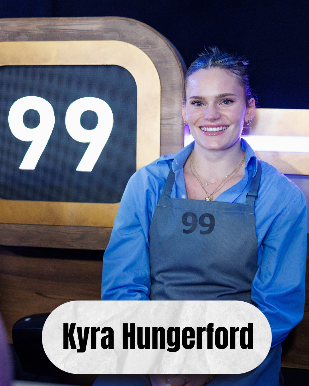 A woman with a bright smile standing in front of a sign with the number 99, wearing a gray apron with the number 99 and a blue shirt, with her name Kyra Hungerford displayed.