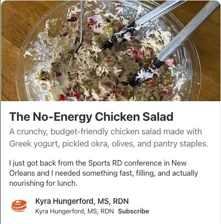 A clear glass bowl containing chicken salad with Greek yogurt, pickled okra, olives, and other ingredients, with two metal utensils in the bowl.