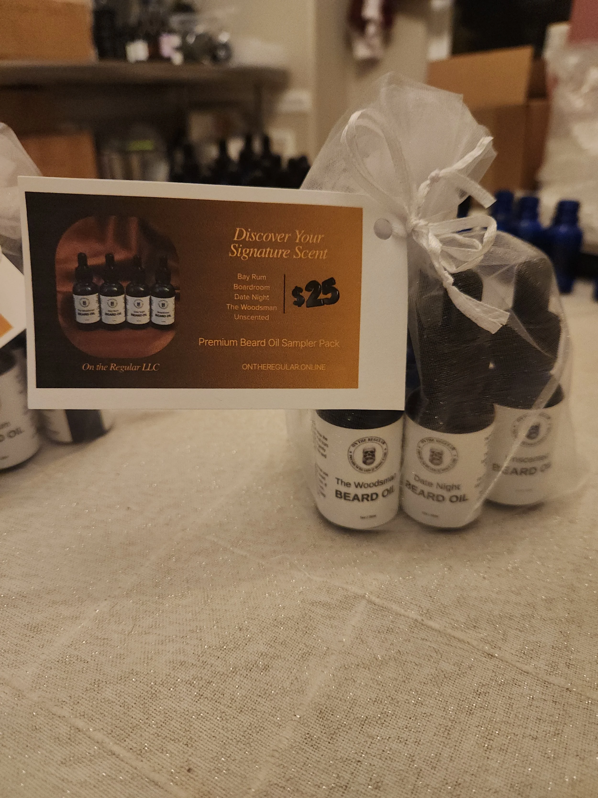 A pack of beard oil bottles and a card promoting a beard oil sampler pack for $25, displayed on a beige tablecloth.