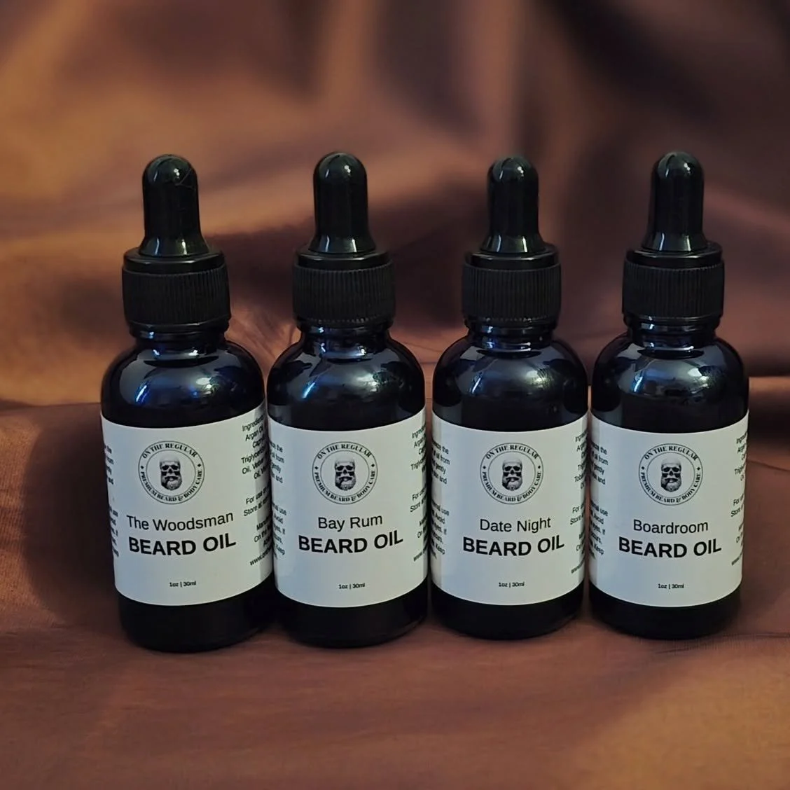 Four bottles of beard oil with black labels and dropper caps, labeled with different scents: The Woodsman, Bay Rum, Date Night, and Boardroom, placed on a wooden surface.