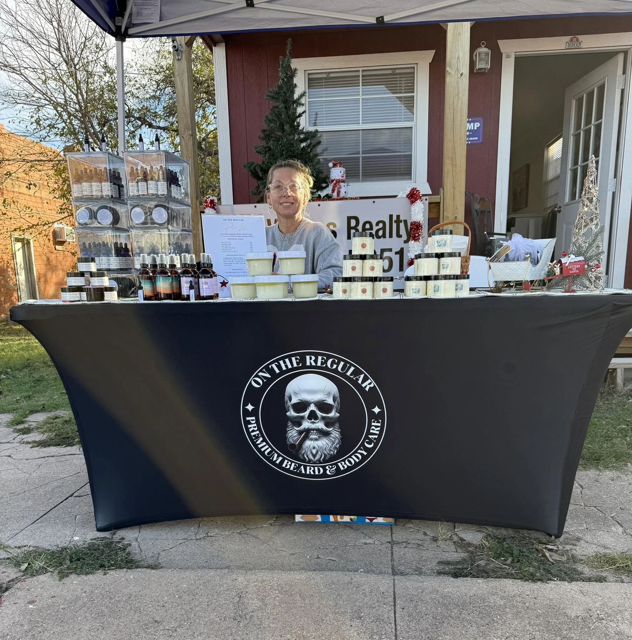 A woman sitting at a booth selling oils at an event. The booth has a black tablecloth with a large skull logo and text, and is decorated with small Christmas ornaments and a small Christmas tree. The woman is smiling and wearing glasses, with a menu and products displayed in front of her. In the background, there is a house with red and white siding, a window, and a door.