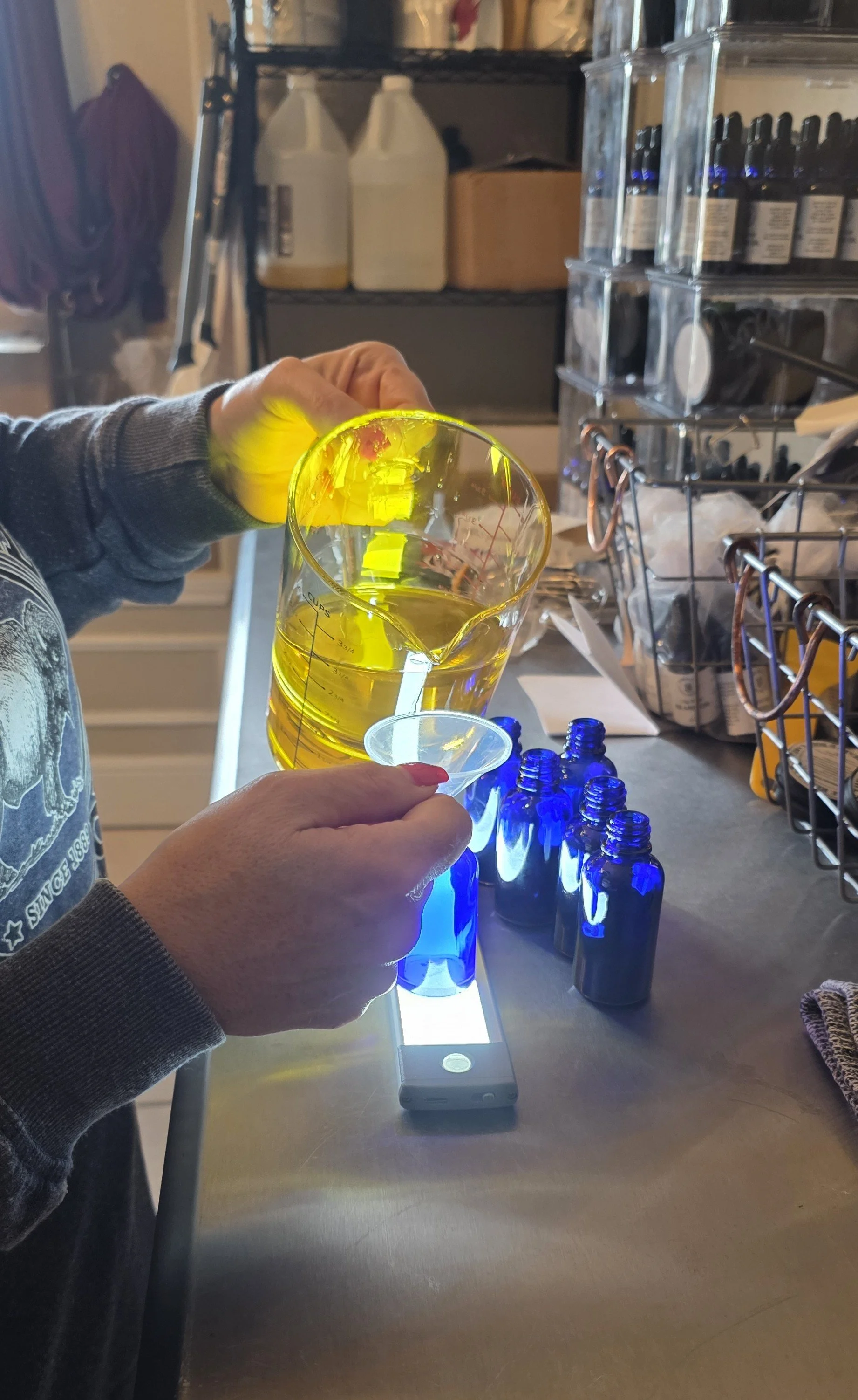Person pouring yellow liquid into small cup at a lab or workshop setting with bottles, containers, and equipment on shelves and table.