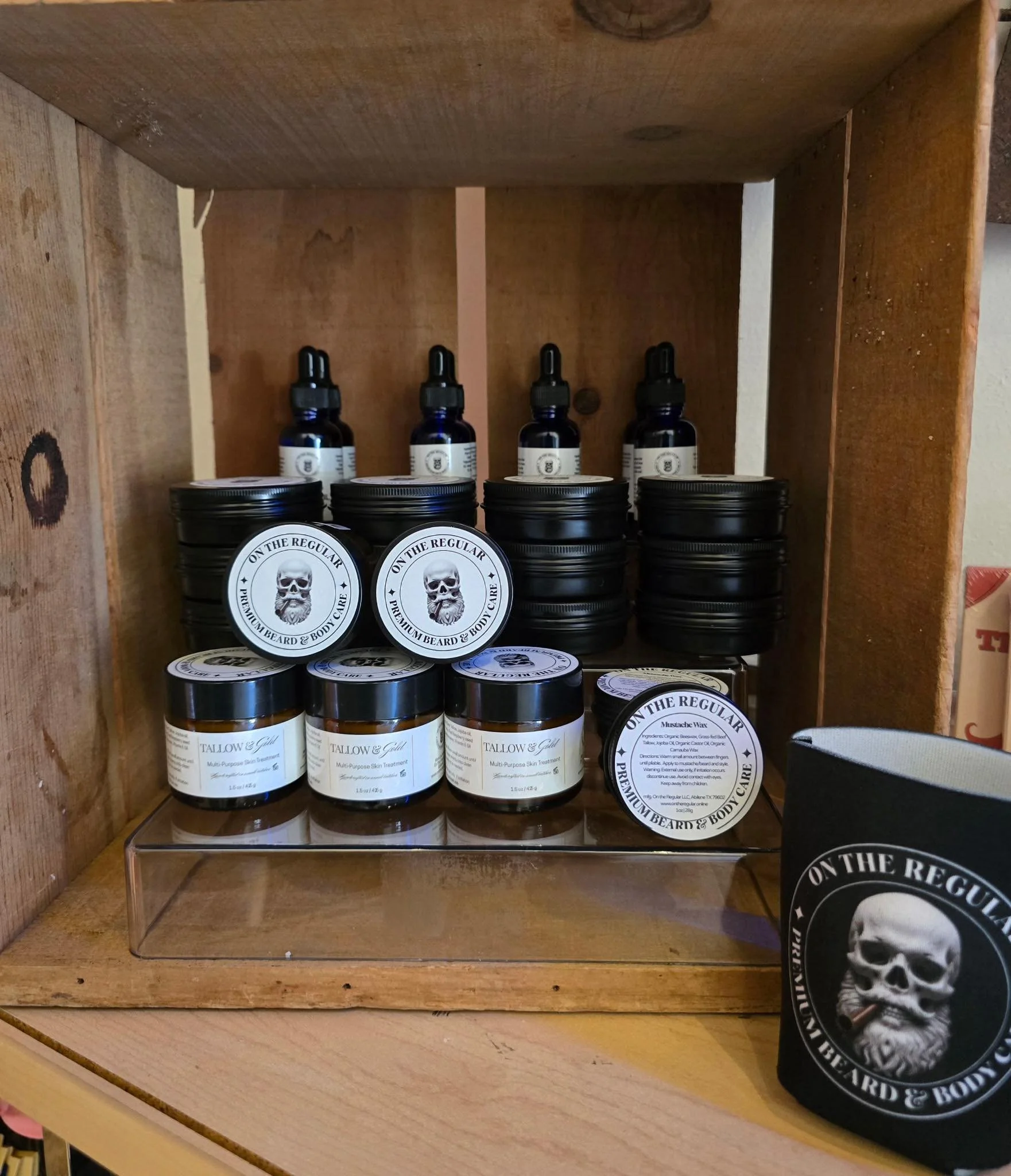 Shelf with grooming and skincare products, including beard balm and mustache wax, in black containers with a skull logo and text, displayed in a wooden cabinet.