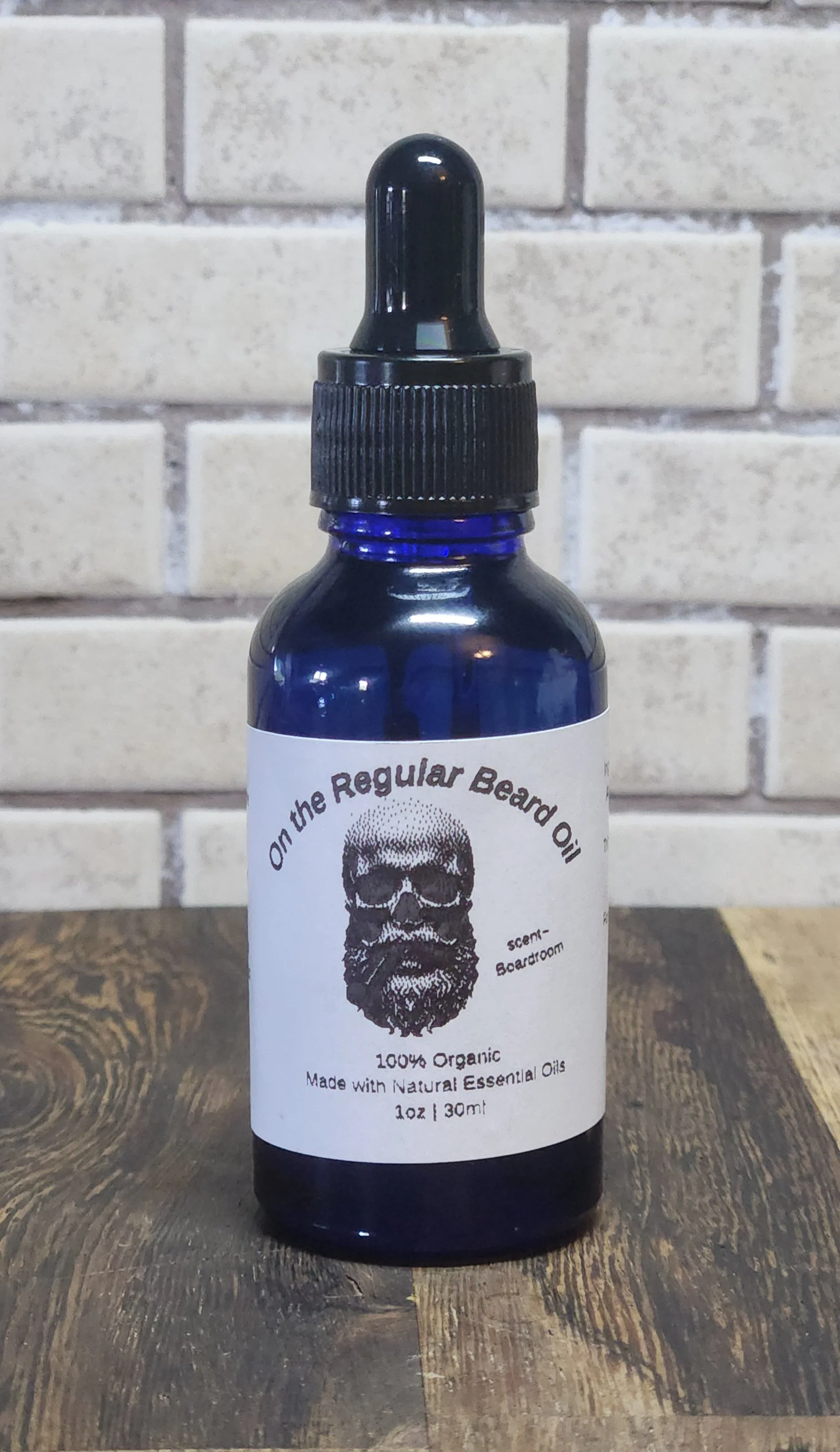 A blue glass bottle with a black dropper cap on a wooden surface, featuring a label with a skull illustration and text about beard oil.
