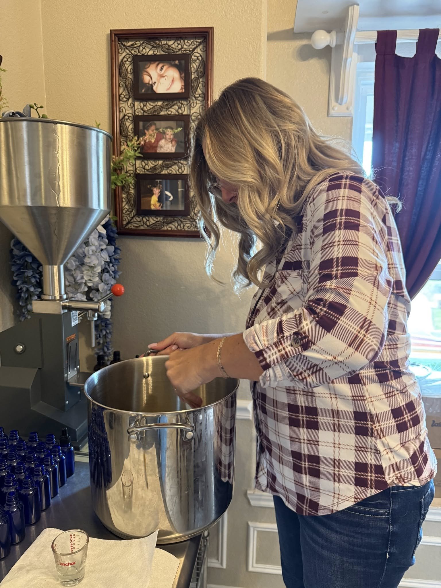A woman with blonde hair wearing glasses, a plaid shirt, and jeans is standing by a stainless steel , stirring or checking its contents. Behind her, there is a window with purple curtains, a wall with a photo collage frame, and some bottles on a table.