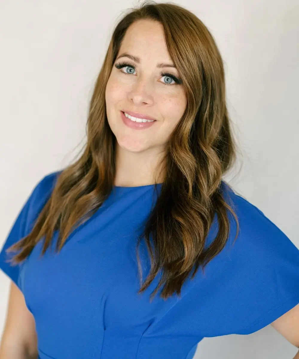 A woman with long, wavy brown hair, blue eyes, and light skin, smiling and wearing a blue top against a plain white background.