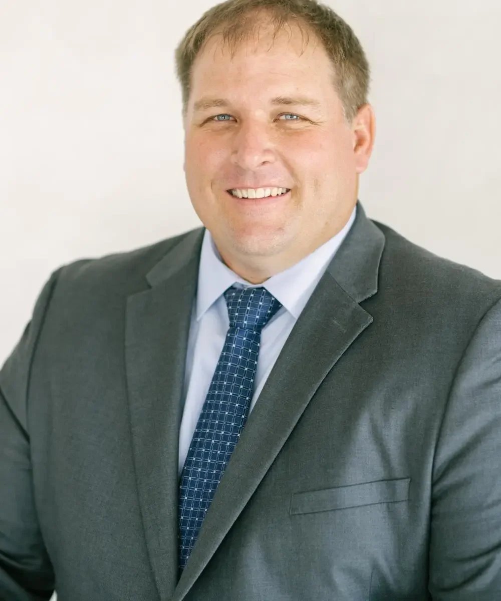 A man in a gray suit with a blue checkered tie, smiling, against a plain light background.