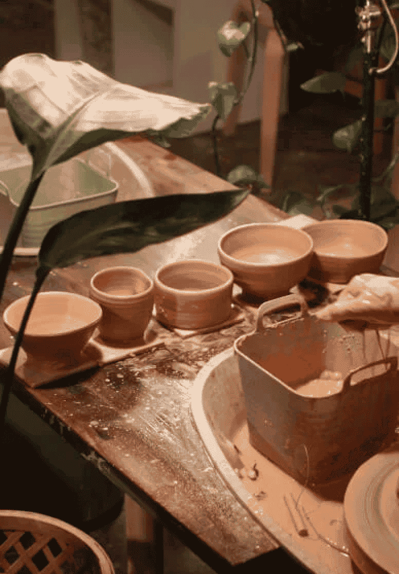 A pottery workspace with several unfinished clay bowls and a rectangular clay piece on a wooden work table, surrounded by pottery tools and a plant leaf in the foreground.