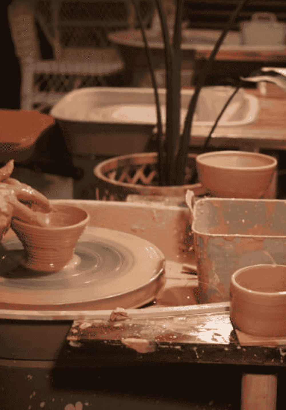 A pottery wheel in use with clay forming a small bowl, surrounded by various pottery tools and other clay items in a dimly lit pottery studio.