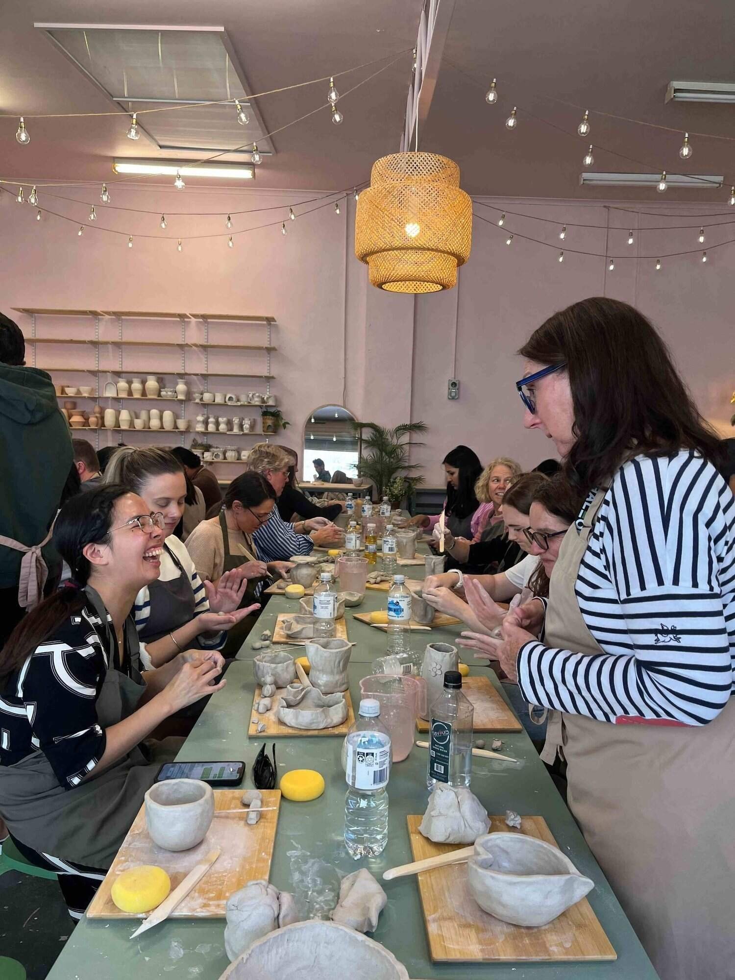 People gathered around a table participating in a pottery workshop, working with clay and pottery tools in a well-lit room with hanging string lights and ceramic ware on shelves in the background.