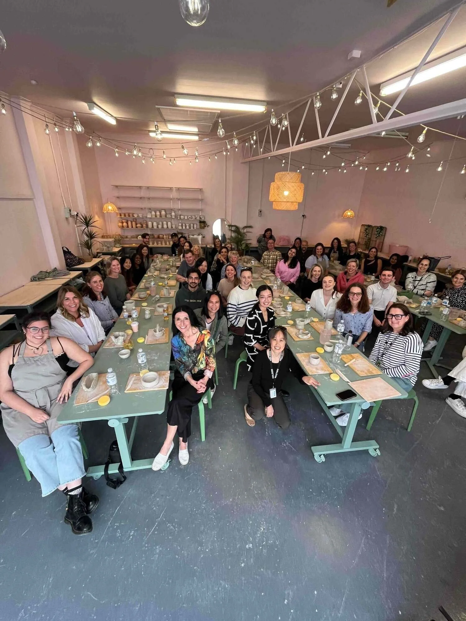 Group of people gathered around a long table in a well-lit room, smiling for a photo, with tables set with plates, cups, and water bottles, and decorated with hanging lights and plants.