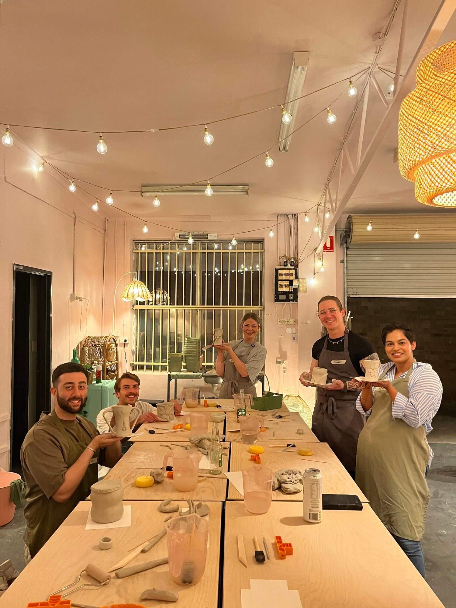 Group of five people in a dining setting, with some holding ceramic cups, smiling, under string lights, with pottery tools on the table, in an indoor space with pink walls.
