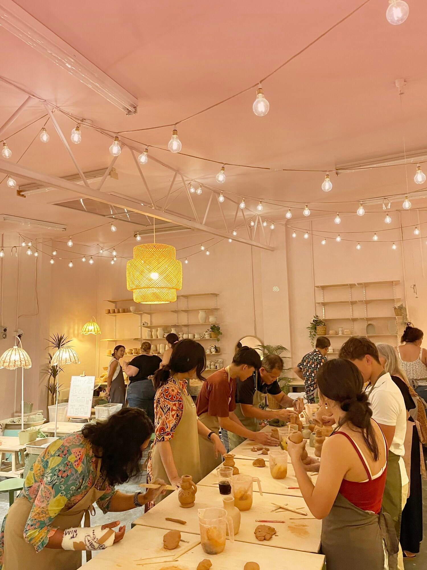 People participating in a pottery workshop at a cafe or studio, working on clay projects at a large table, with shelves of pottery and plants in the background, illuminated by hanging and table lamps.