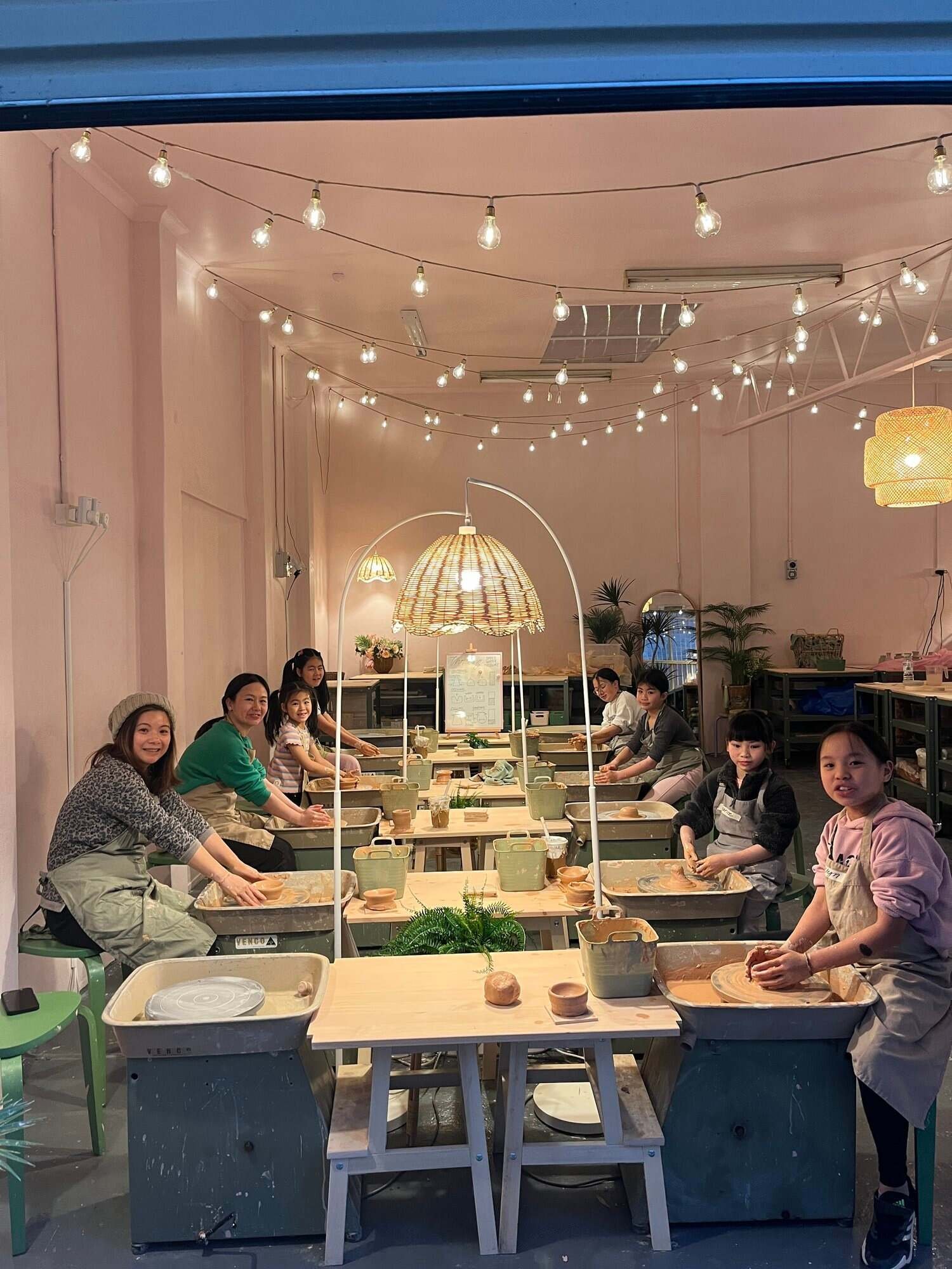 Group of people at pottery workshop, working with clay on pottery wheels, indoors with pink walls and hanging string lights.